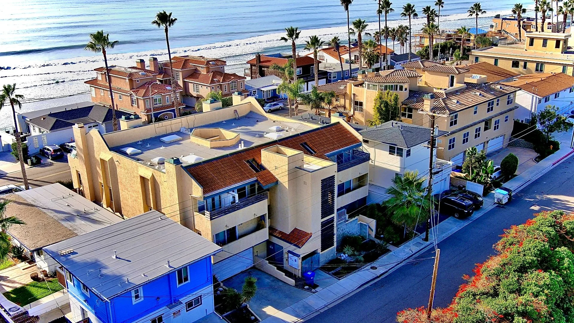 Aerial view of a coastal neighborhood with beach houses, palm trees, and the ocean in the background. The scene includes residential buildings with tiled roofs, parked cars, and vibrant greenery.