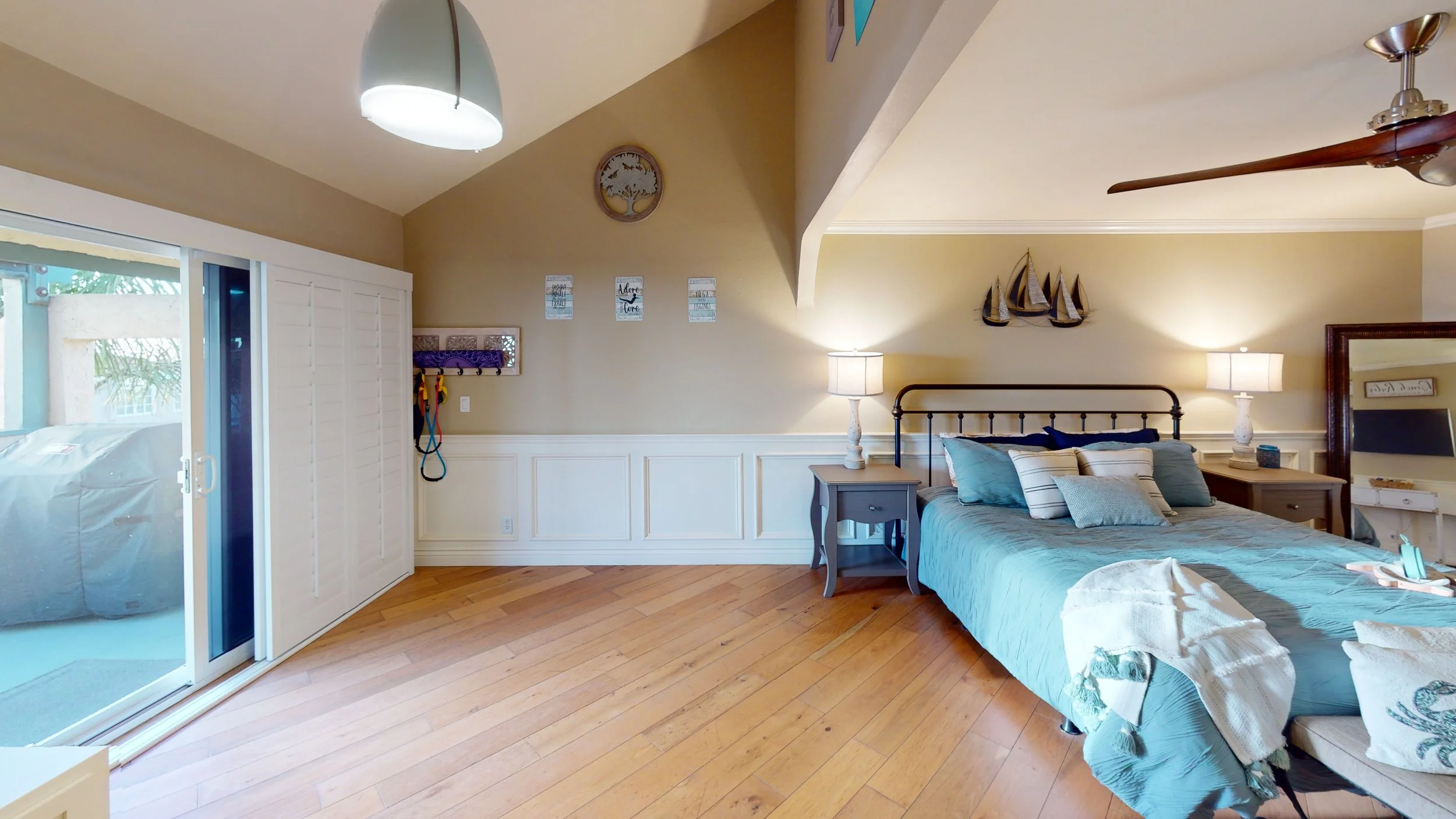 A bedroom with a metal bed frame, blue bedding, and multiple pillows. Two nightstands with lamps sit on either side of the bed. A sailboat wall decoration hangs above the headboard. There's a sliding glass door with white plantation shutters leading 