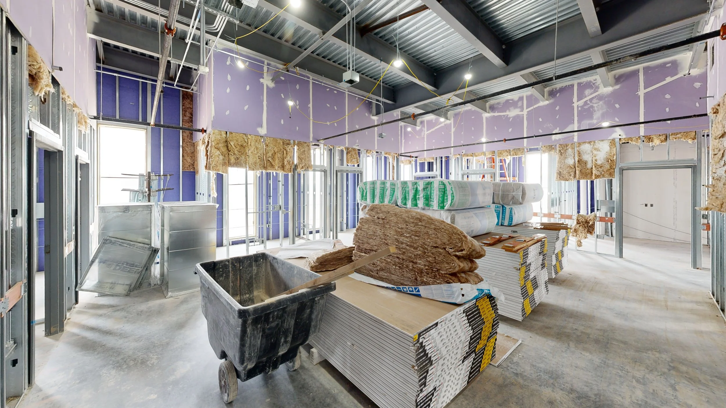 Interior of a building under construction with exposed metal framing, insulation, drywall in progress, and construction materials like drywall sheets, insulation rolls, and a wheelbarrow with construction debris.
