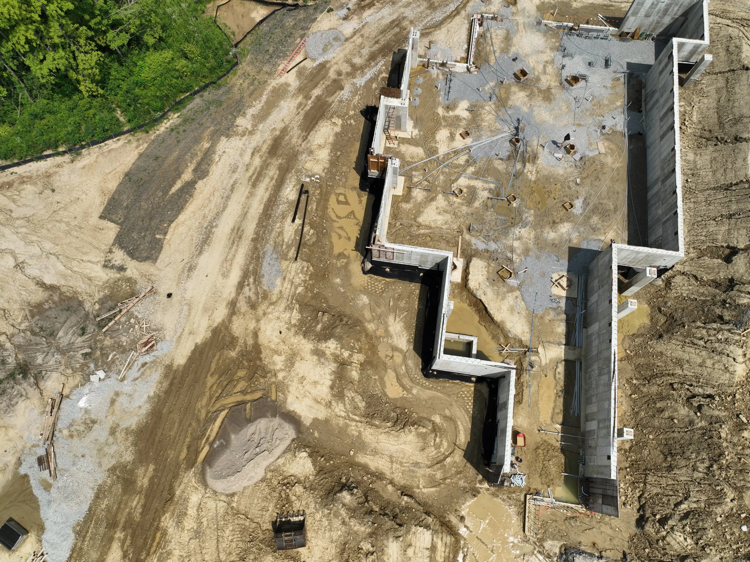 Aerial view of a construction site with partially built concrete walls, dirt roads, and construction materials, surrounded by dirt and gravel terrain.