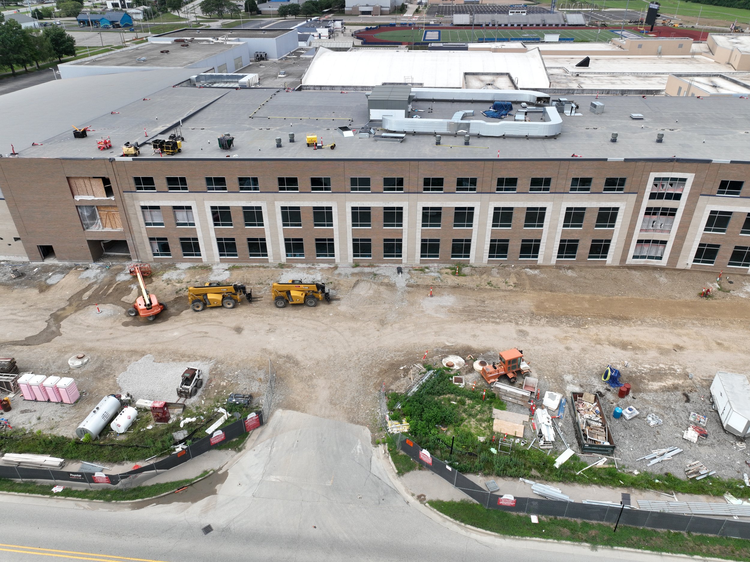 Construction site with building in progress, construction equipment, and trucks on dirt lot in front of the building. Visible parking lot and athletic field in the background.