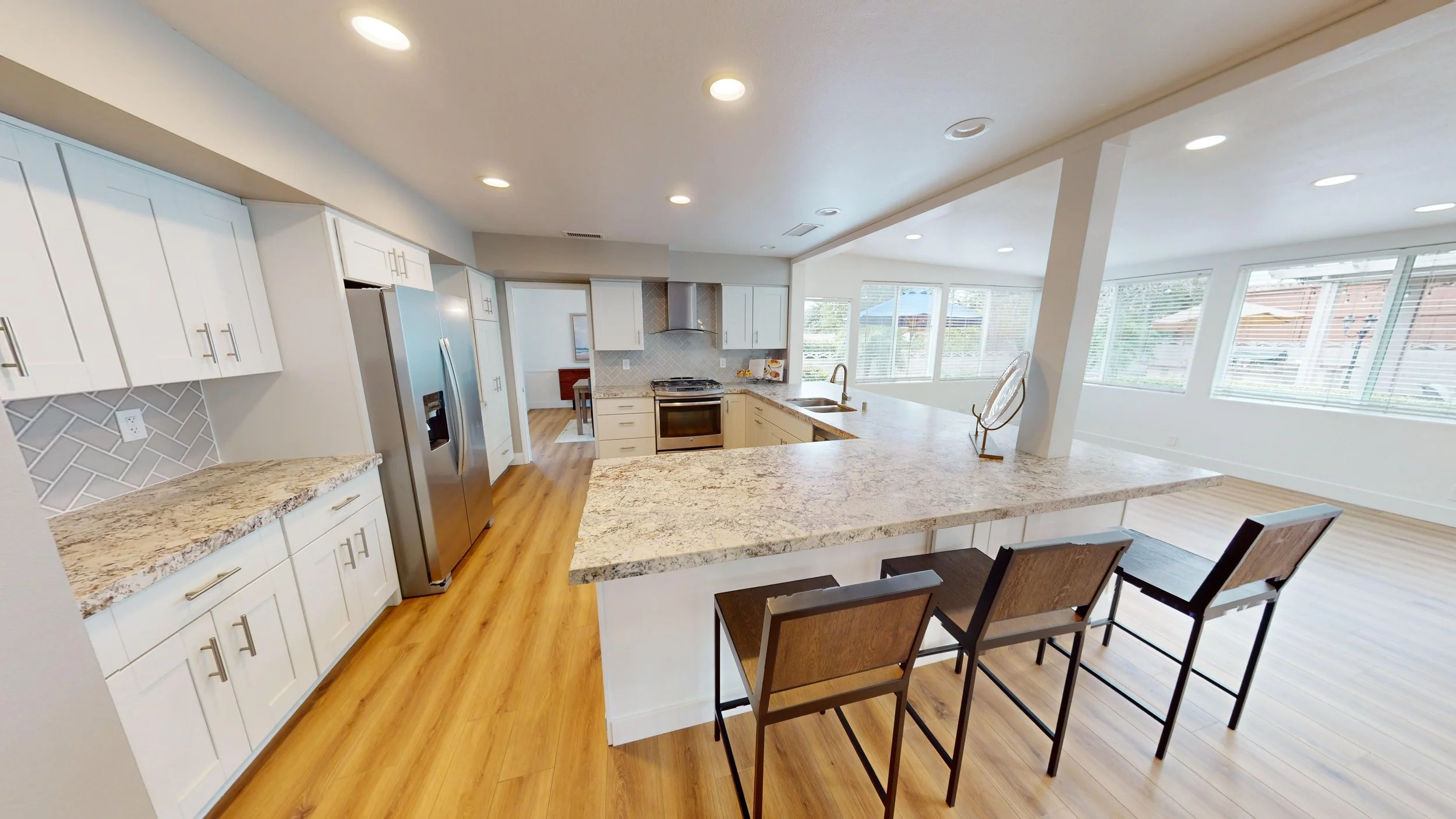 Open-concept kitchen with white cabinets, stainless steel refrigerator, granite countertops, and a breakfast bar with three chairs, adjacent to a bright living area with large windows.
