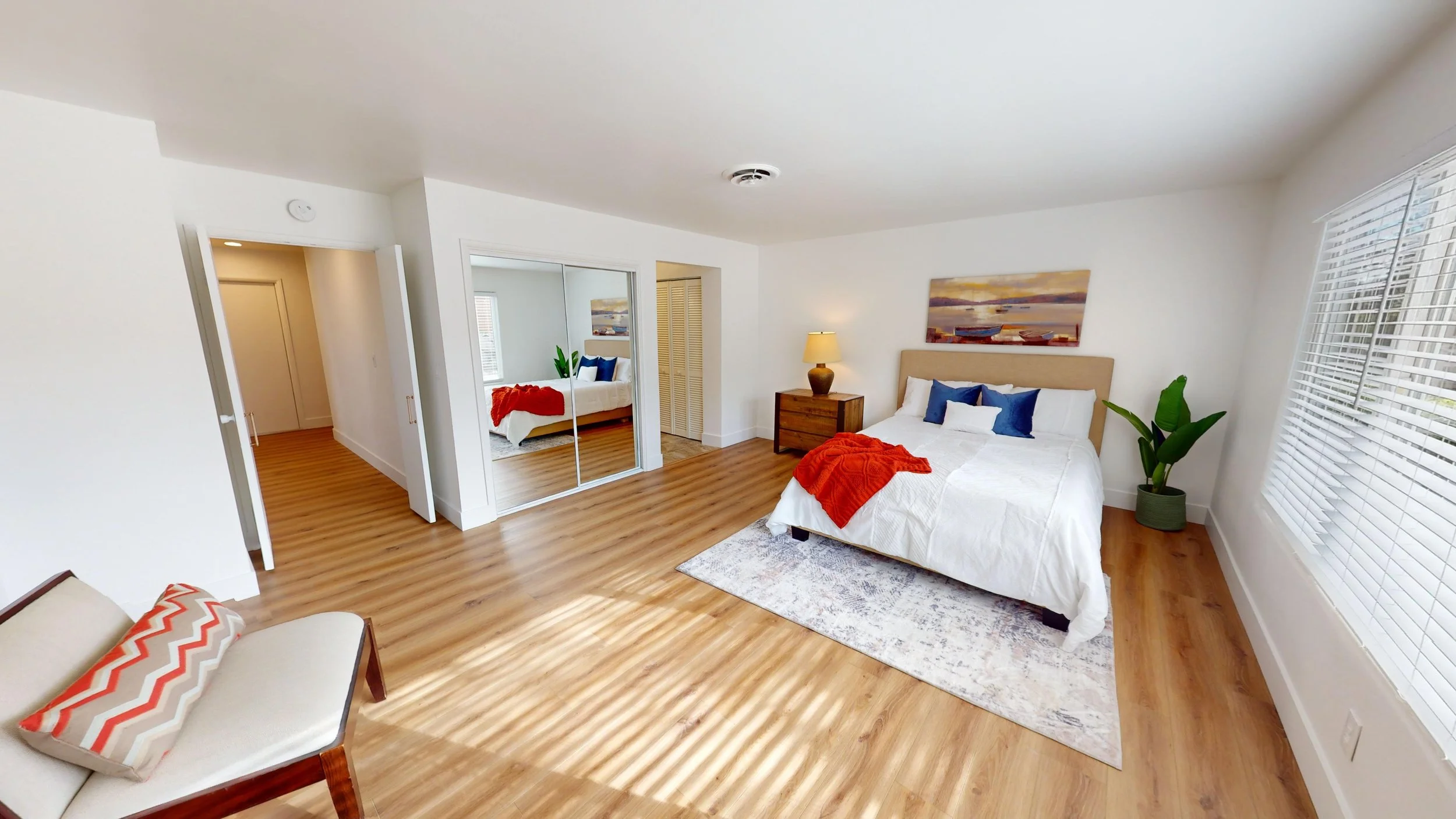 A spacious bedroom with a large bed, white bedding, blue pillows, and an orange blanket, next to a wooden nightstand with a lamp. There is a large window with blinds on the right, a potted plant, and a piece of artwork above the bed. A mirror closet,