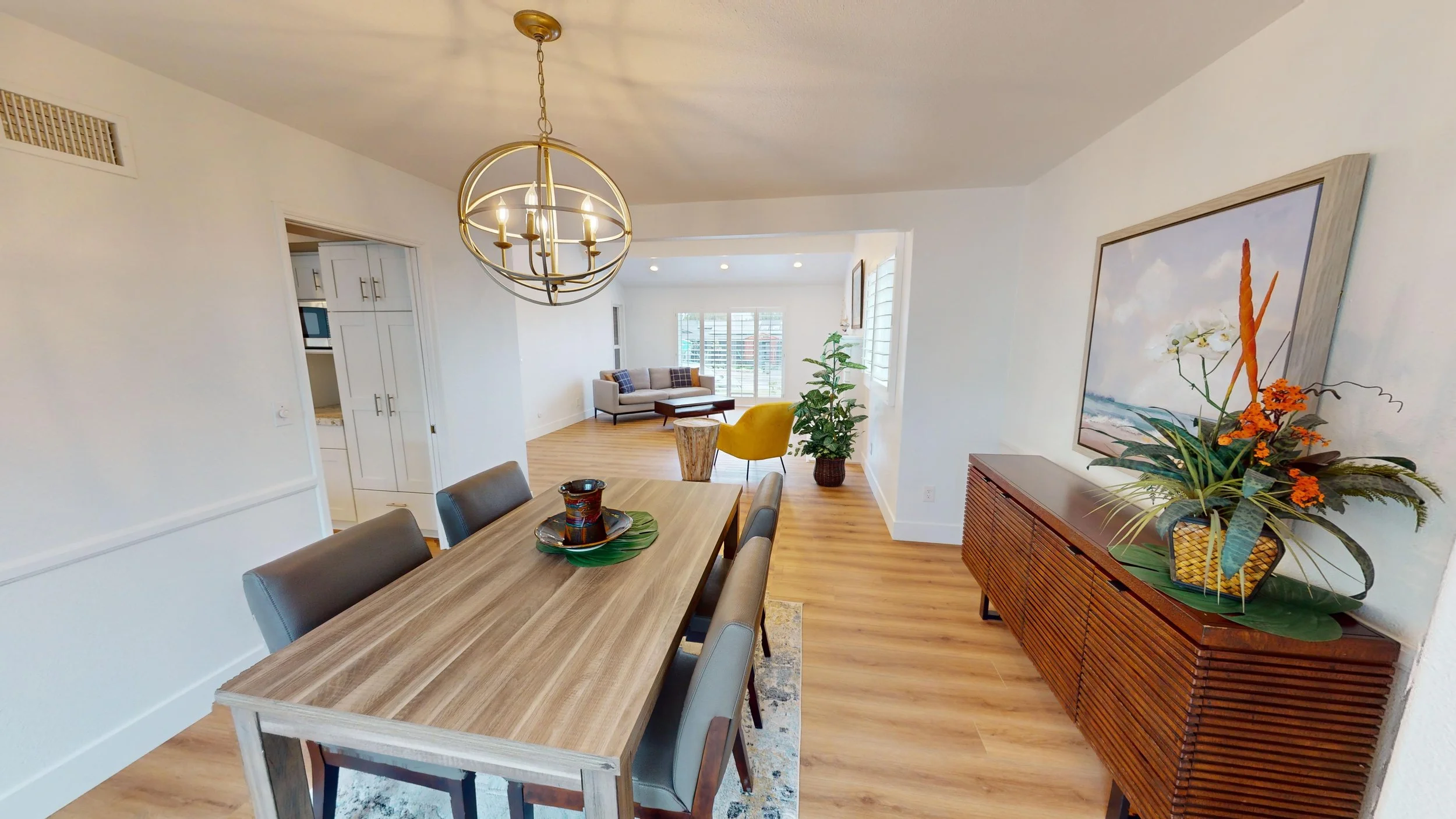 Interior of a modern living and dining room with a wooden dining table, chairs, artwork, a sideboard with a floral arrangement, and a sitting area with a sofa, armchair, and large windows.