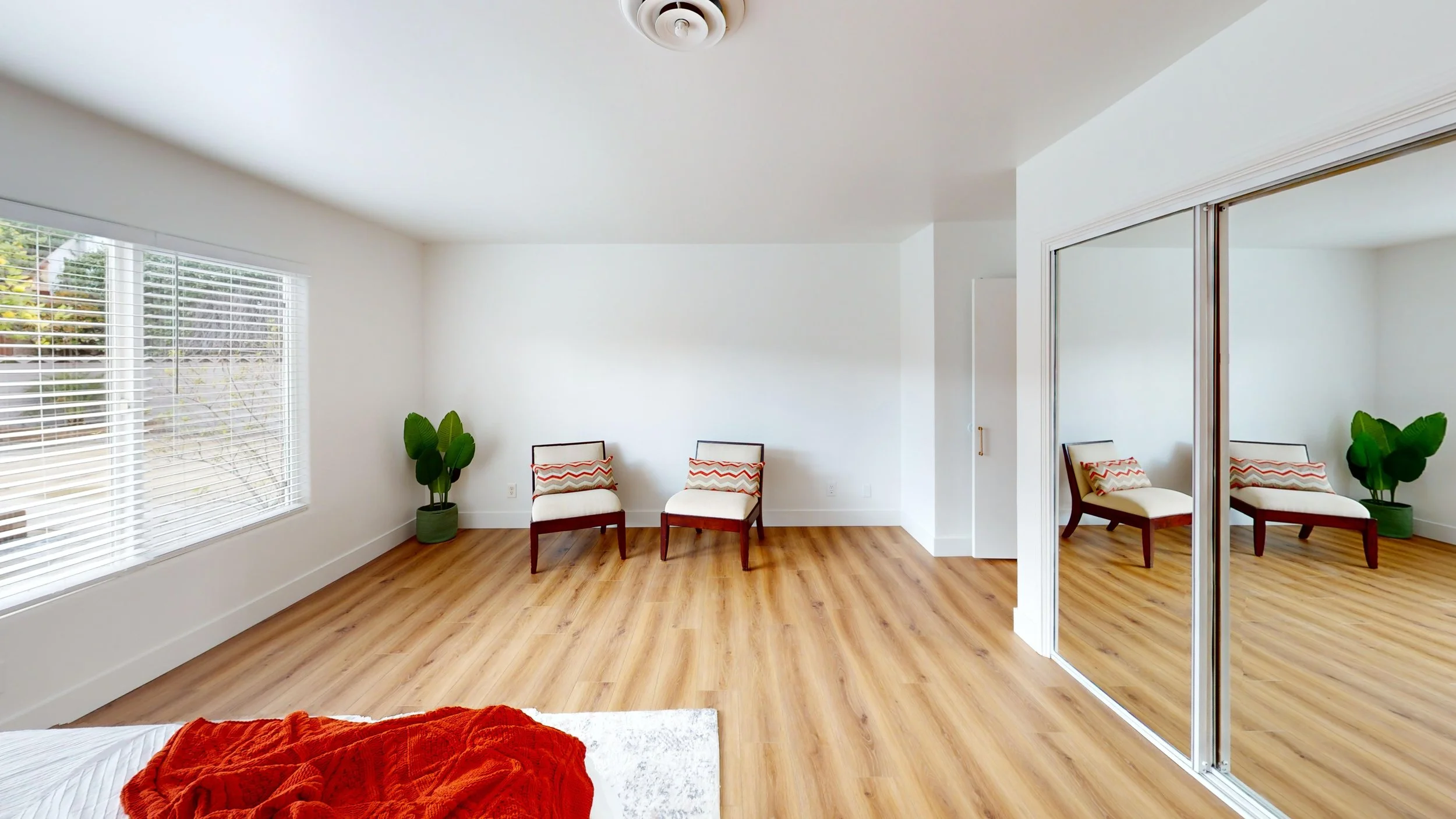 Bedroom with light wood flooring, white walls, two chairs with patterned pillows, a large window with blinds, a mirrored closet door, and a potted plant near the chairs.
