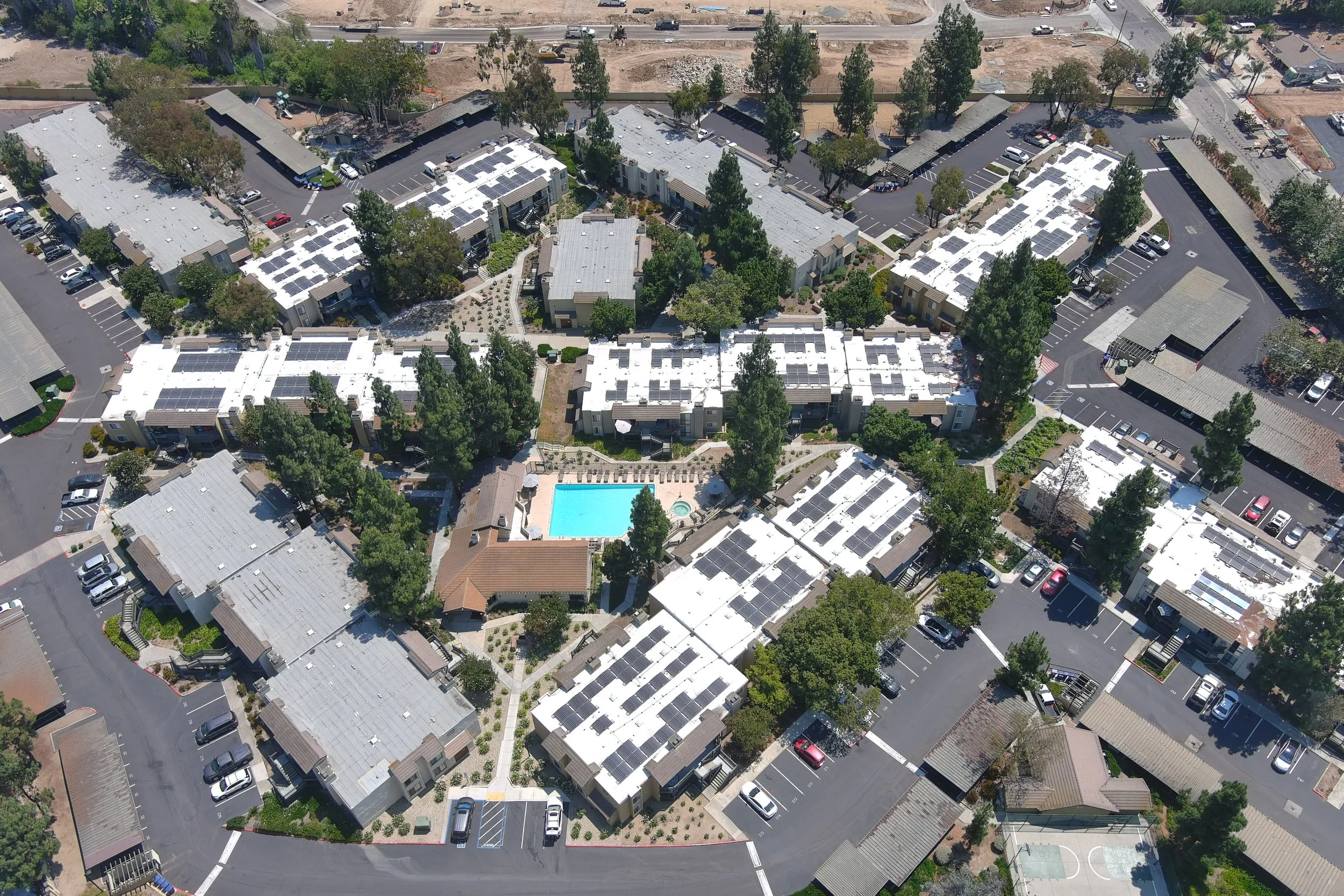 Aerial view of a residential apartment complex with multiple buildings, a large swimming pool in the center, surrounded by trees, parking lots, and walkways.