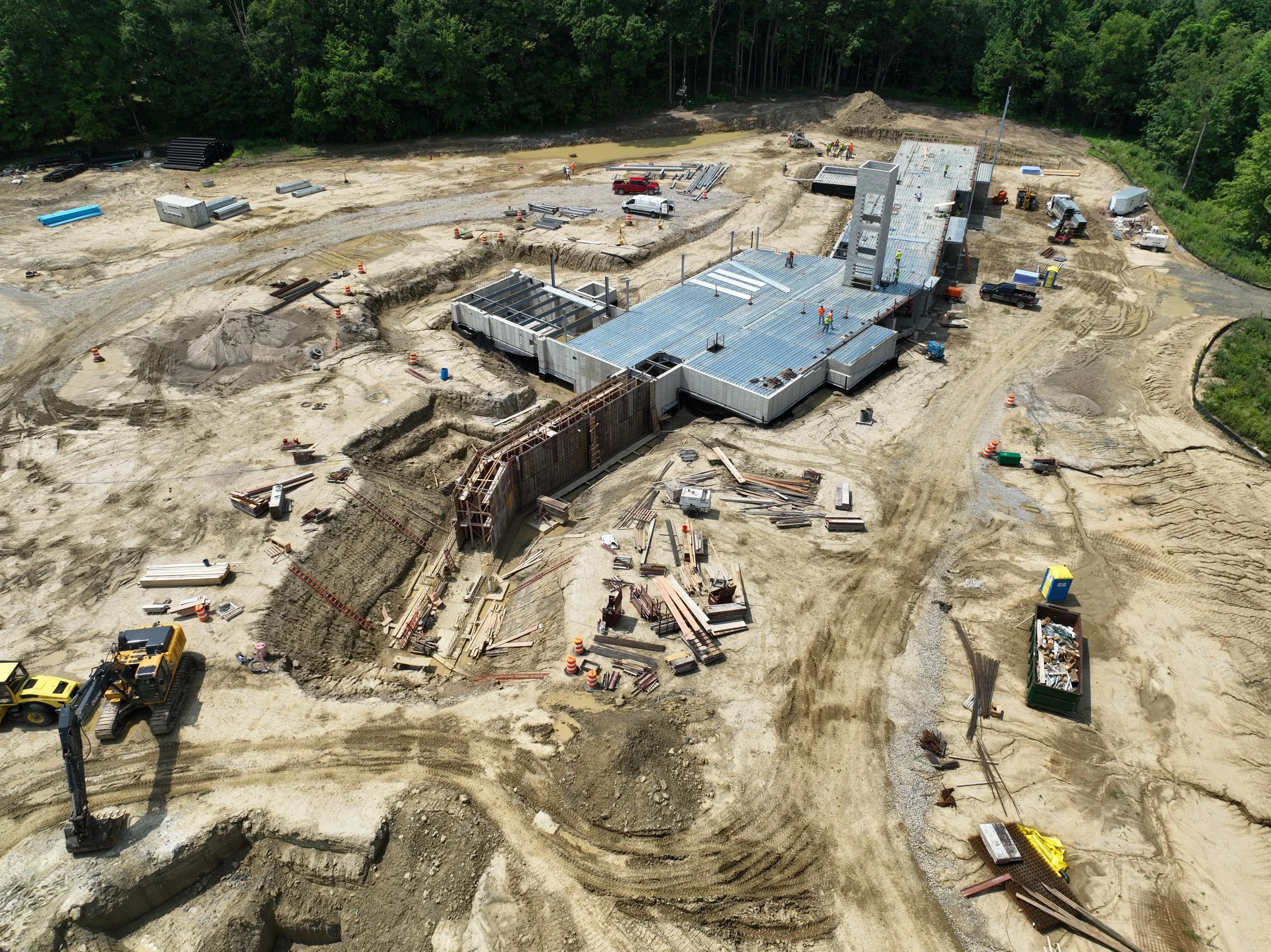 Aerial view of a construction site with a partially built structure, construction workers, machinery, and construction materials scattered across the dirt area, surrounded by trees in the background.