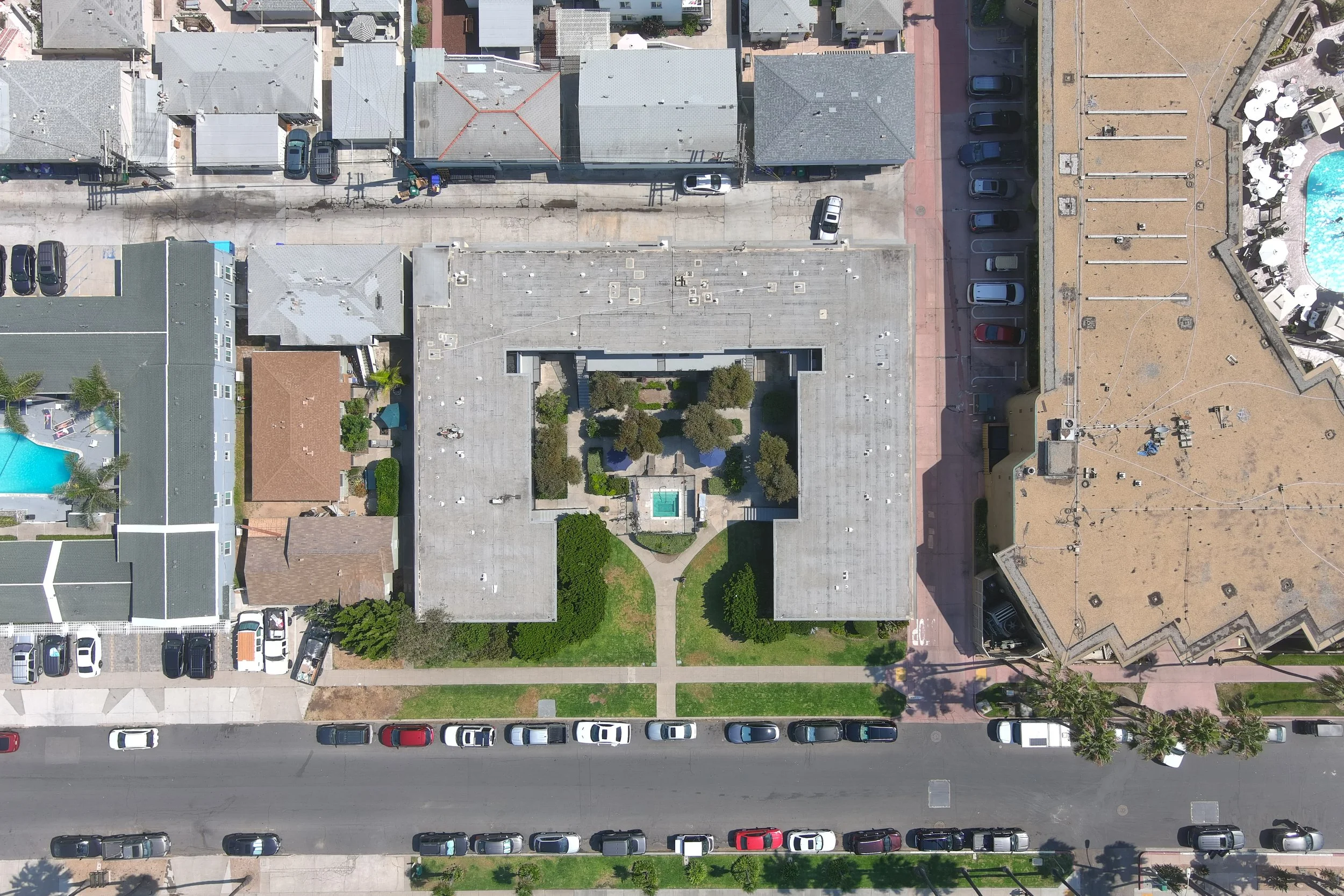 Aerial view of a city block showing a central courtyard with green grass, trees, and a small fountain, surrounded by buildings and parking lots.