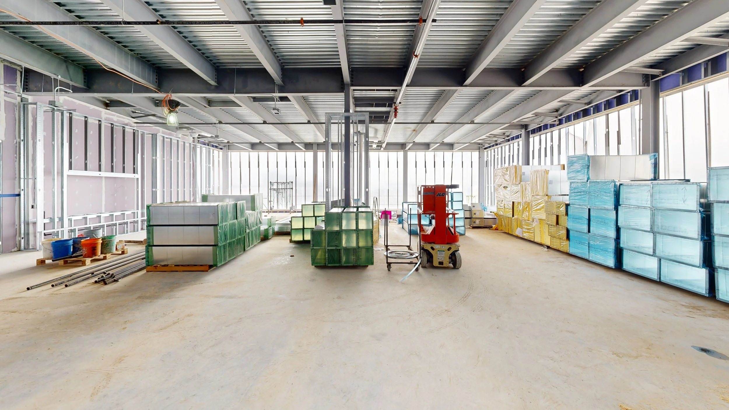 Interior of a building under construction with metal framework, stacks of glass or acrylic panels, construction materials, and equipment.