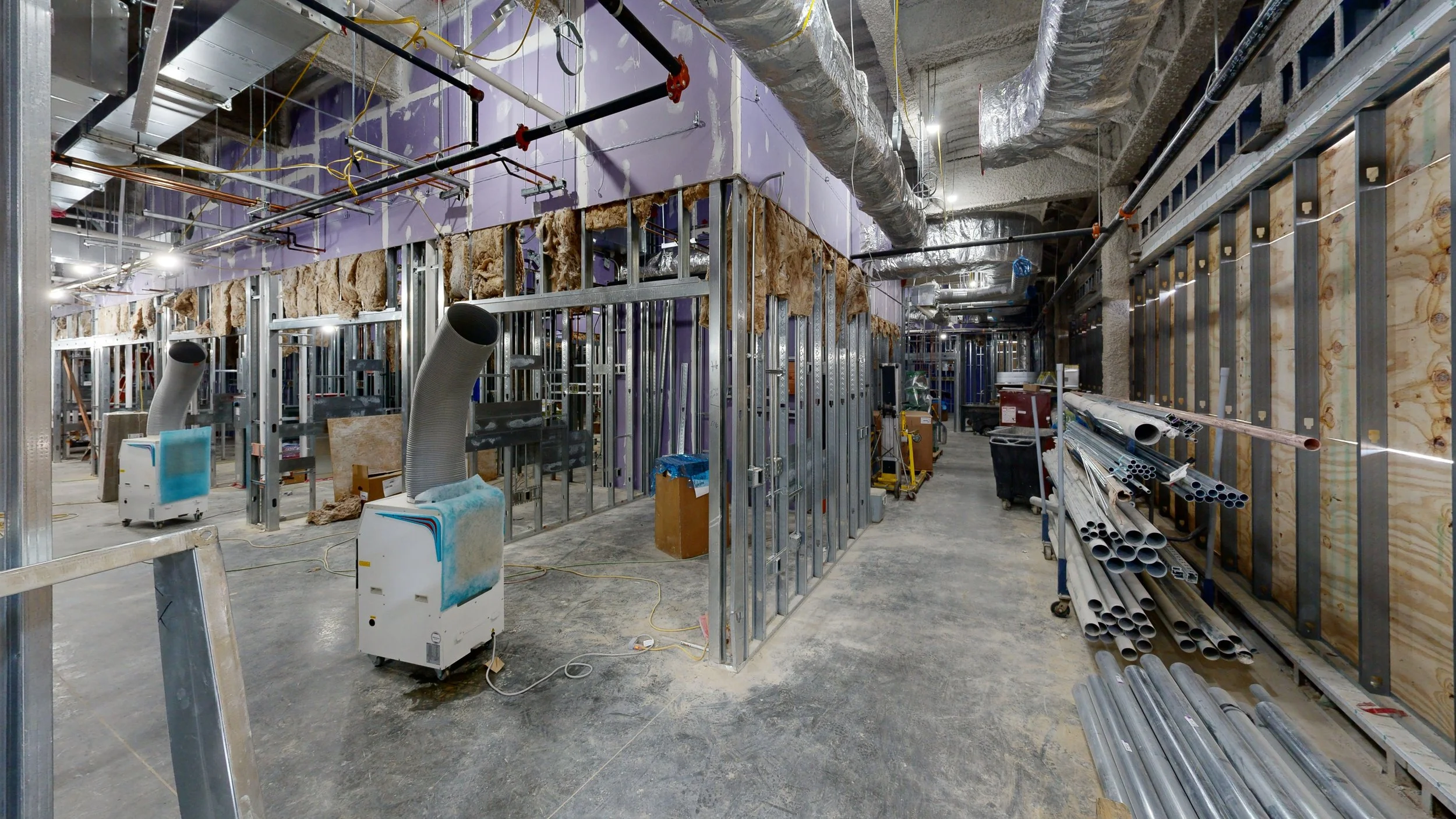 Construction site with metal framing and exposed insulation, pipes, and ducts, indicating ongoing interior building work.
