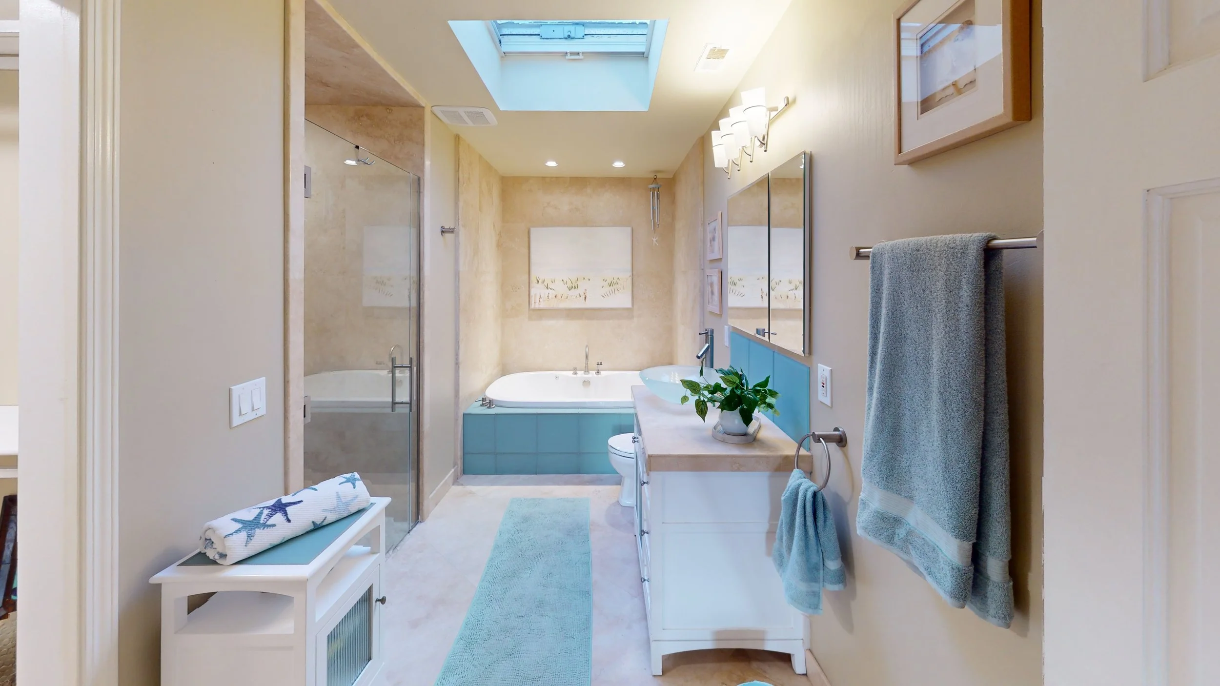 Bright bathroom with a skylight, a soaking tub, a glass walk-in shower, a white vanity with a vessel sink, and blue accents, including towels and tiles.