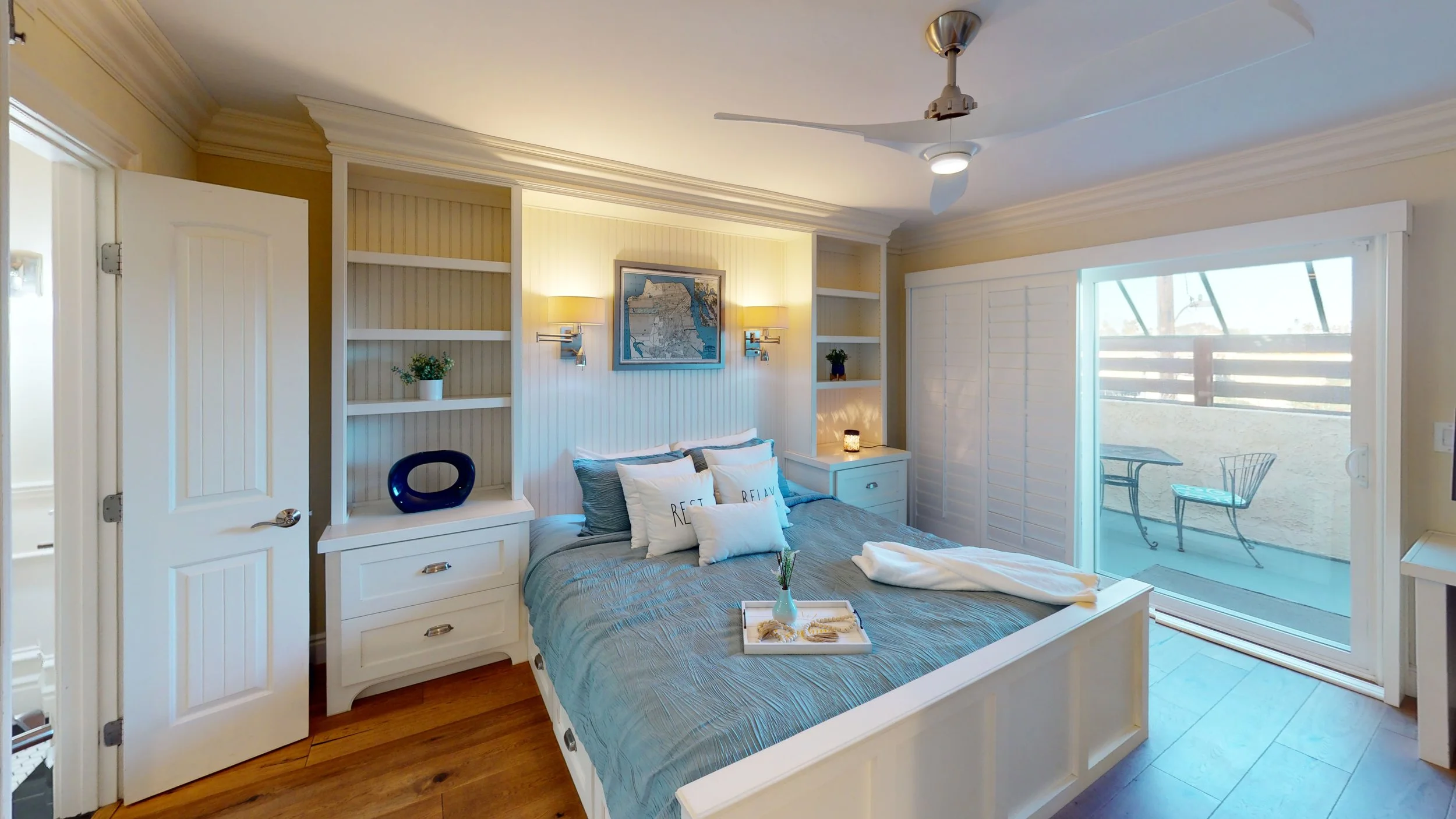 Bedroom with white furniture, pillows reading "REST" and "RELAX," a tray with a vase and seashells on the bed, and sliding glass door leading to a patio with outdoor chairs.