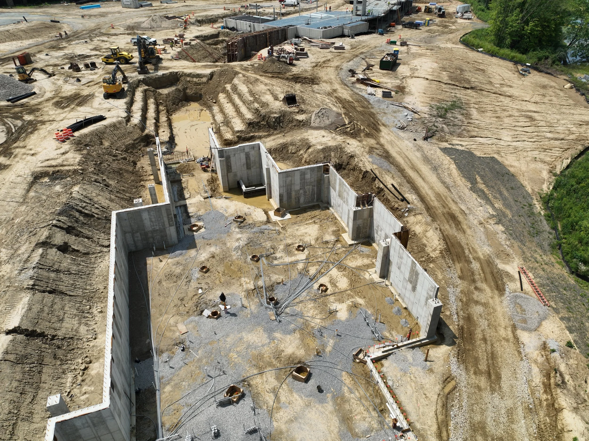 Construction site with partially built concrete foundation walls, construction equipment, unpaved roads, and excavated earth. Some green trees are visible in the background.