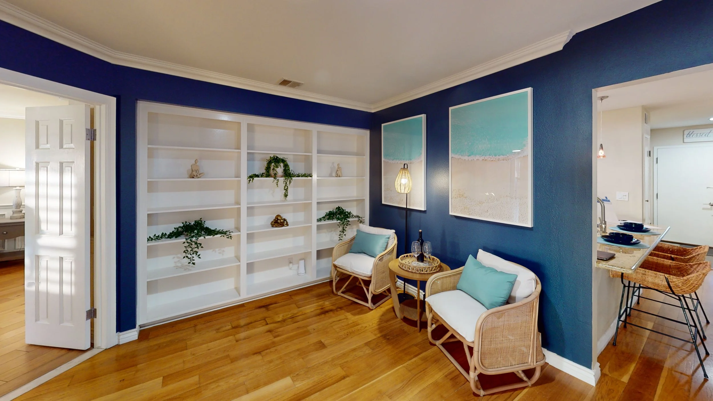 Cozy corner of a living room with two white chairs with teal pillows, a small wooden side table with wine glasses and a bottle, blue accent wall, large beach-themed paintings, and a white bookshelf with decorative items and plants.