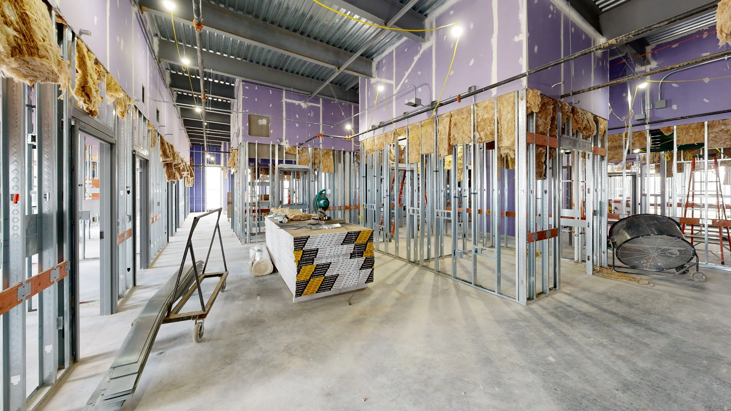 Interior view of a building under construction with exposed metal framing, insulation, and purple drywall on the walls. Construction materials and tools such as a saw, ladder, and rolled insulation are visible.