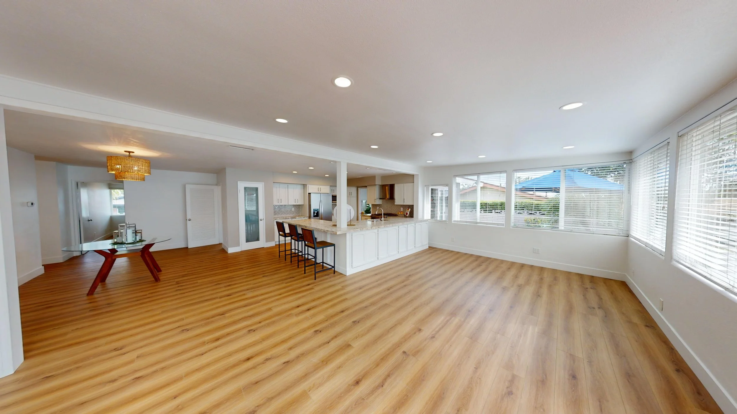 Empty living room with large windowed wall, hardwood floors, and an open kitchen with island and bar stools.