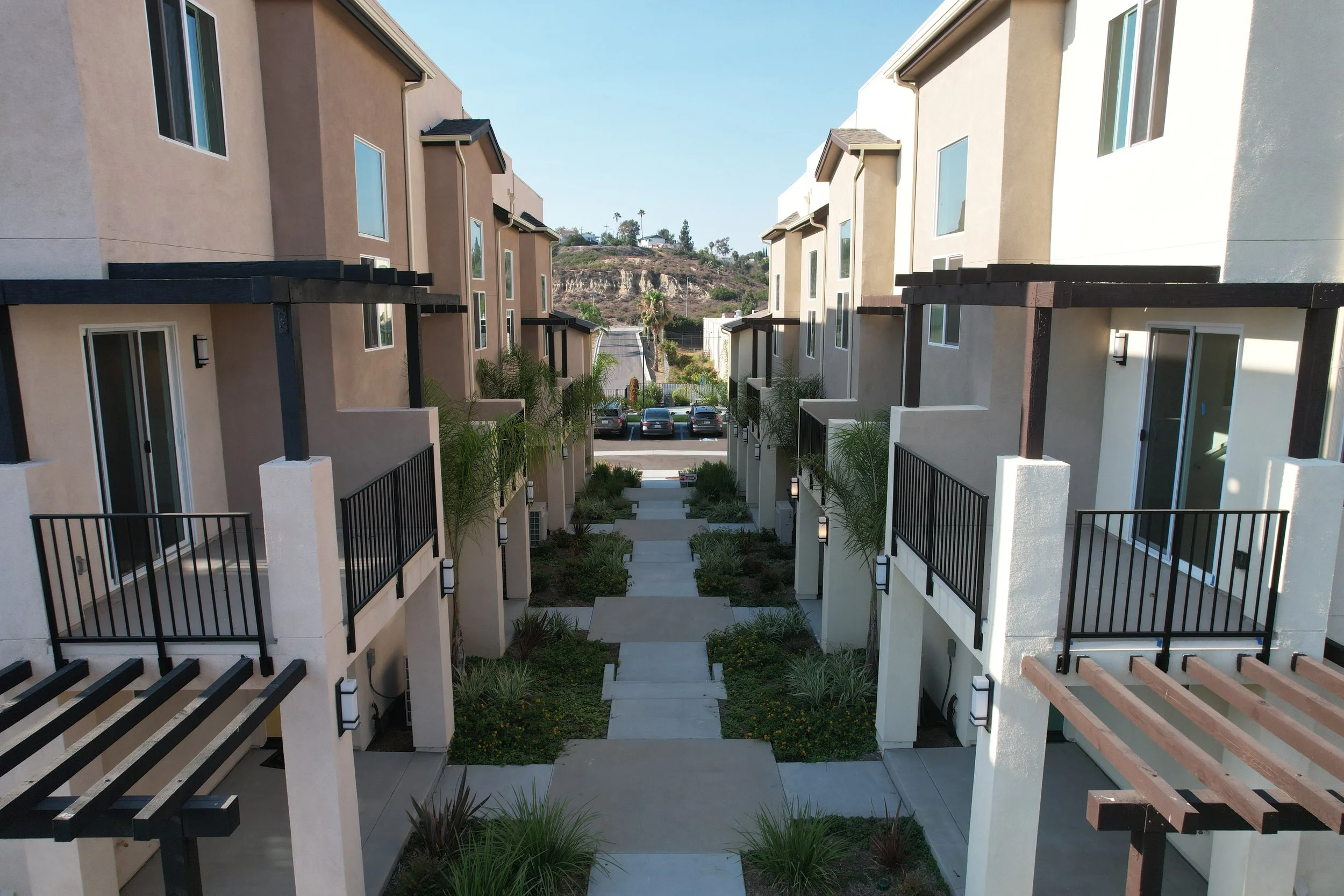 View of a modern apartment complex courtyard with pathways, small landscaped gardens, and two-story buildings with balconies and large windows.