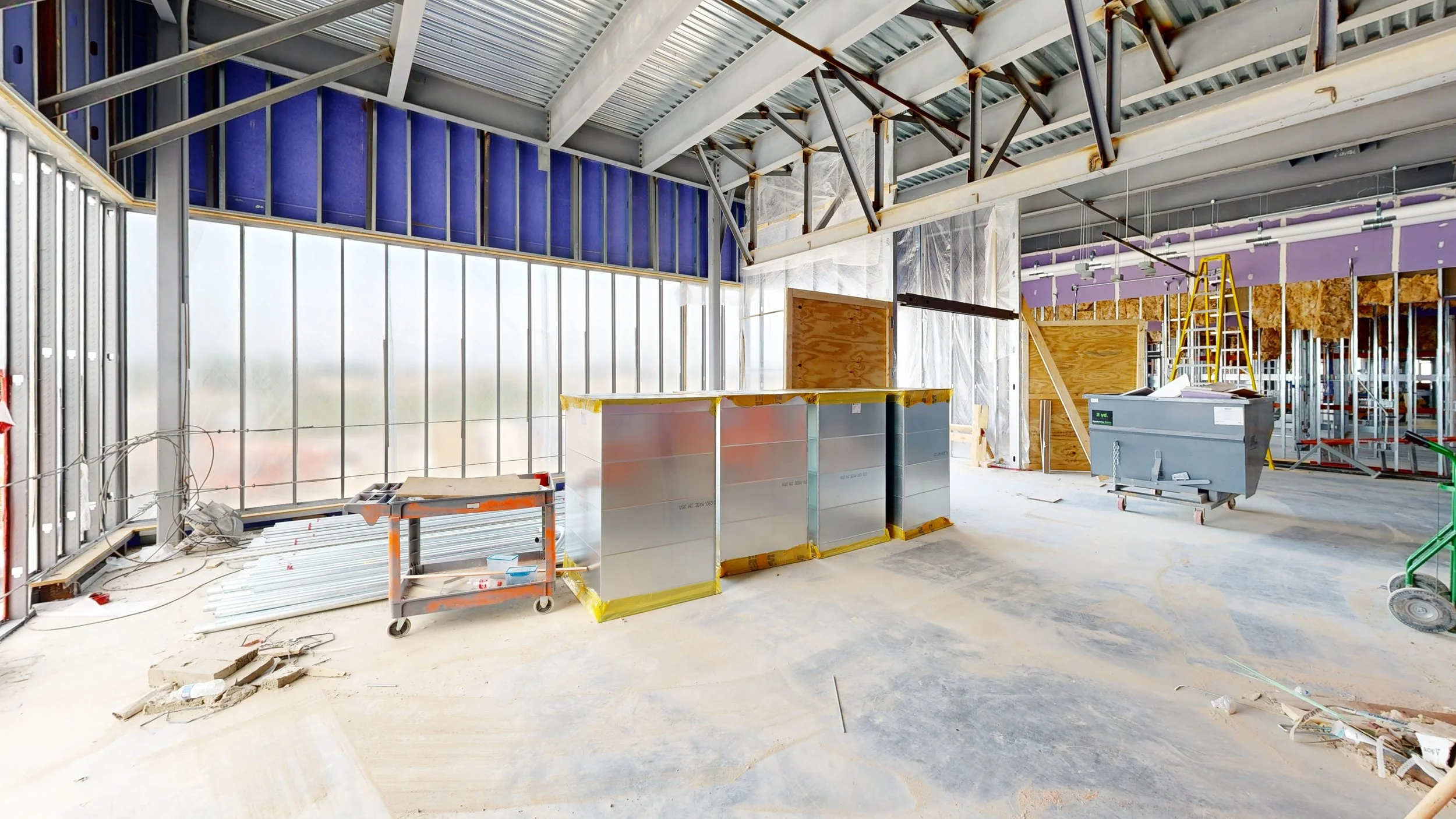 Interior of a building under construction with exposed metal framework, construction materials, and equipment.