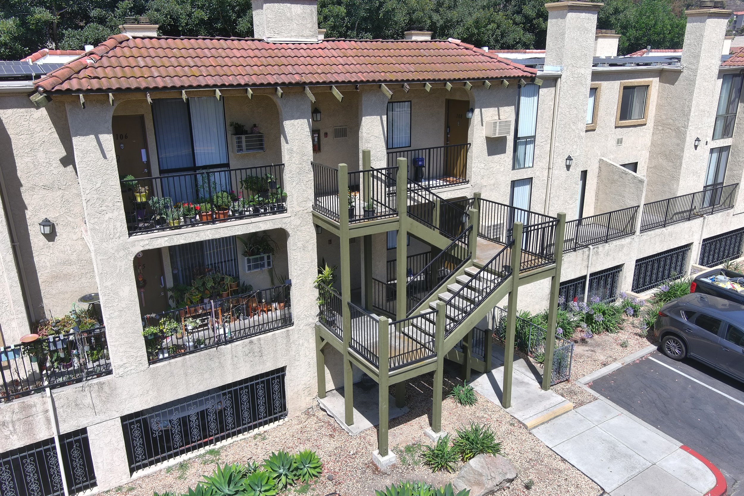 Exterior view of a multi-unit apartment building with balconies, stairs, parking lot, and surrounding landscaping.