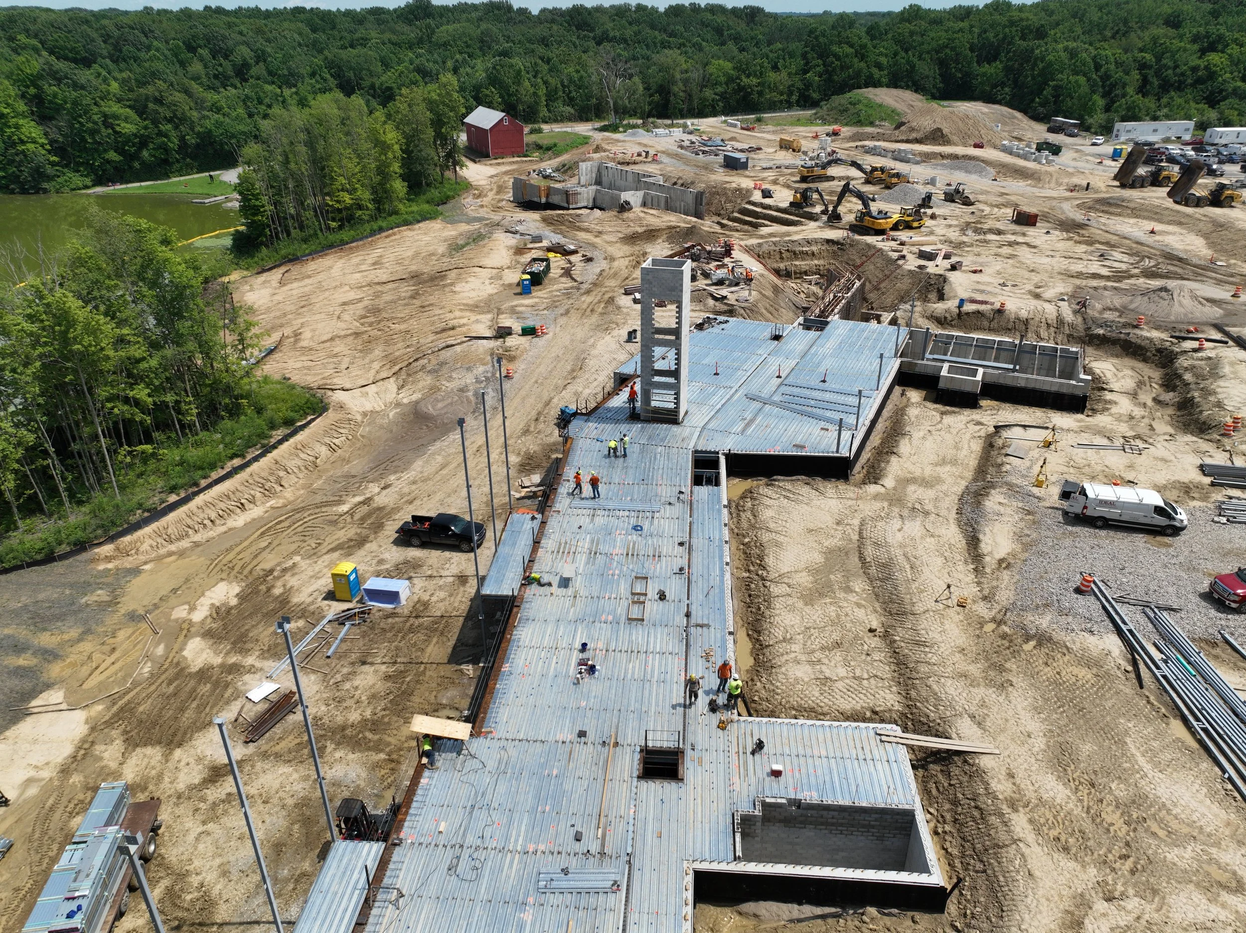 Aerial view of a large construction site where a building is under construction, with workers and equipment visible, surrounded by dirt, gravel, and some trees and a small body of water nearby.