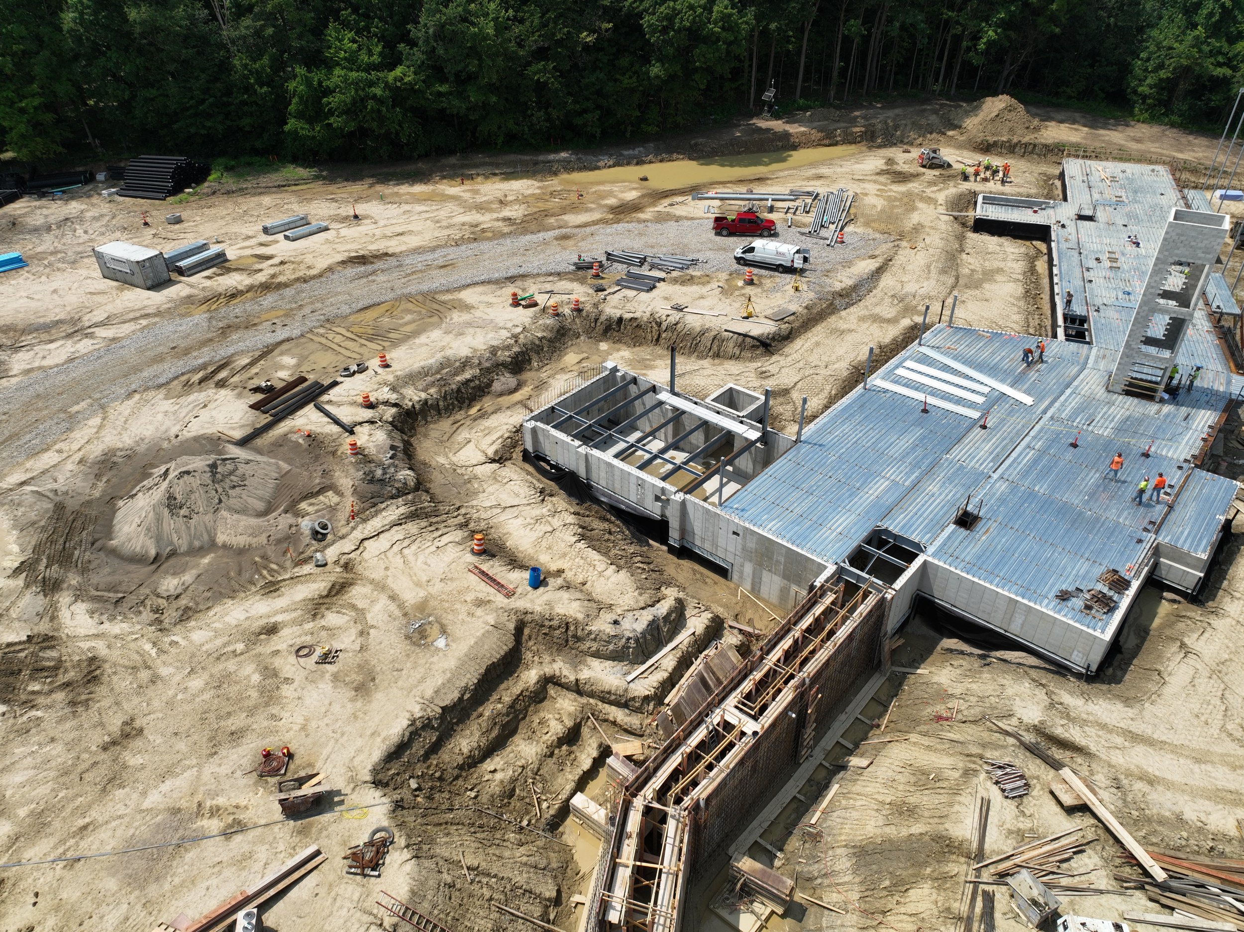 Construction site with building framework, workers, vehicles, and construction materials on dirt ground, surrounded by trees.