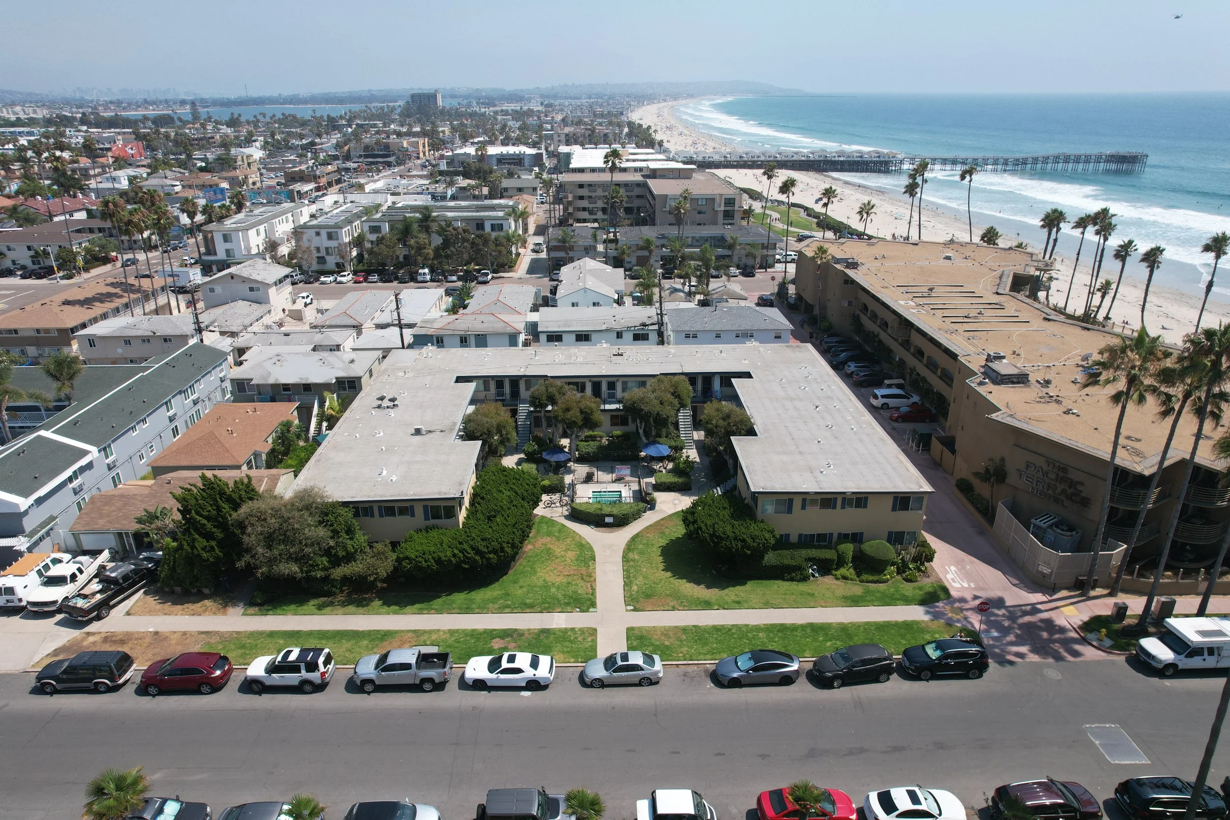 Aerial view of a beachside neighborhood with apartment buildings, palm trees, and a pier extending into the ocean, turquoise water, and sandy beach.