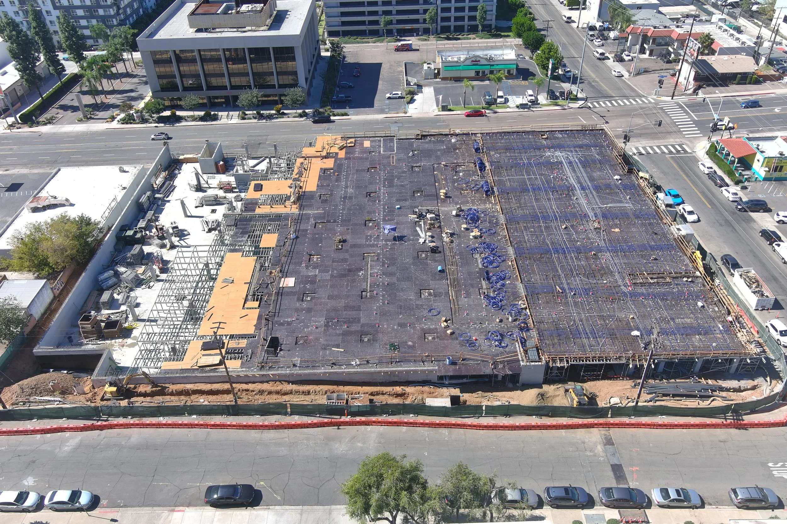 Aerial view of a building under construction with a partially completed roof, construction materials, and equipment visible; surrounding streets with parked cars and nearby buildings.