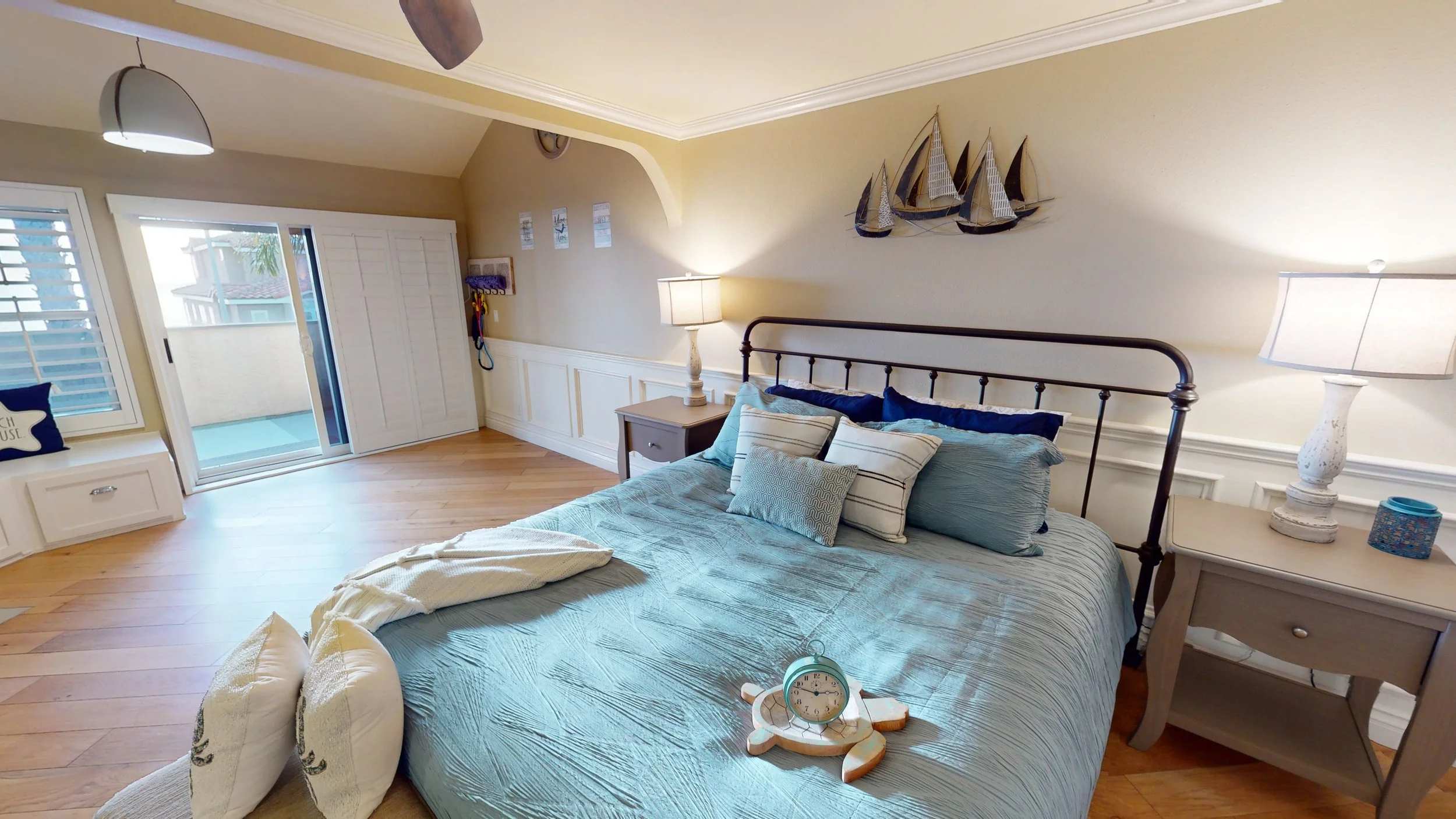 A cozy bedroom with a bed covered in blue bedding and multiple pillows, two nightstands with lamps, a wooden flooring, a sliding door leading to a balcony, and nautical decor on the wall.