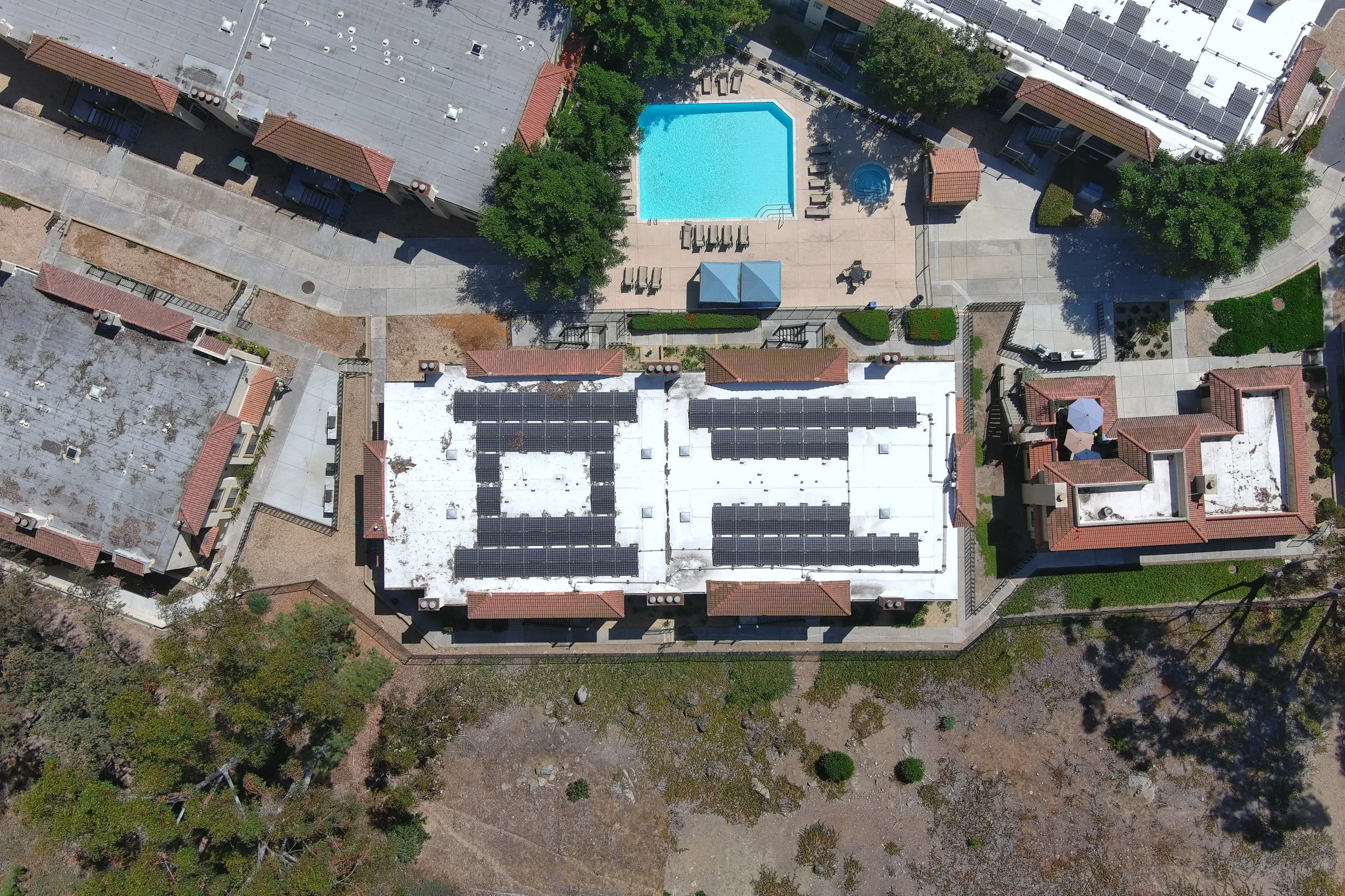 An aerial view of a rooftop with solar panels, a swimming pool, and surrounding buildings and trees.