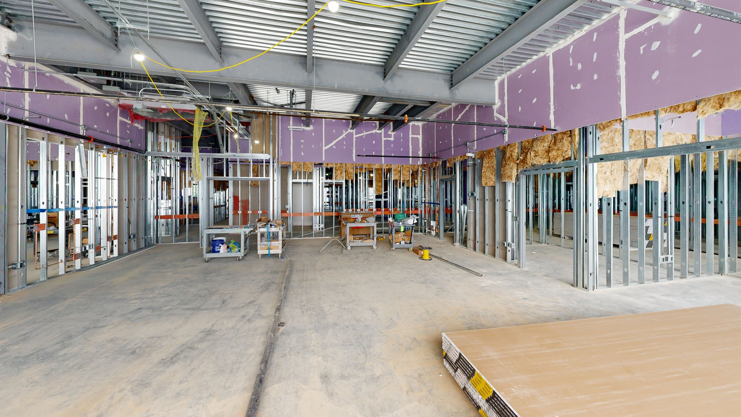 Interior view of a building under construction with exposed steel wall frames, purple insulation boards, and drywall, with construction tools and materials on the floor.