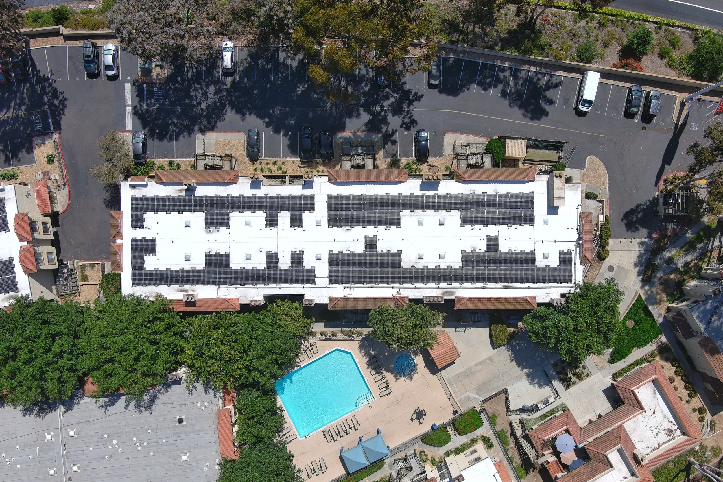 Aerial view of an apartment complex with a large white roof covered in solar panels. There is a parking lot with several cars, a swimming pool, and surrounding trees.