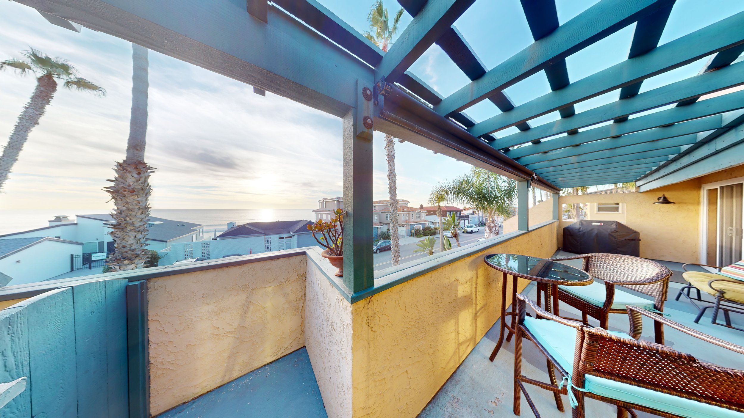 Beachside balcony with outdoor furniture, potted plant, grill, and ocean view at sunset, palm trees, and neighboring houses.