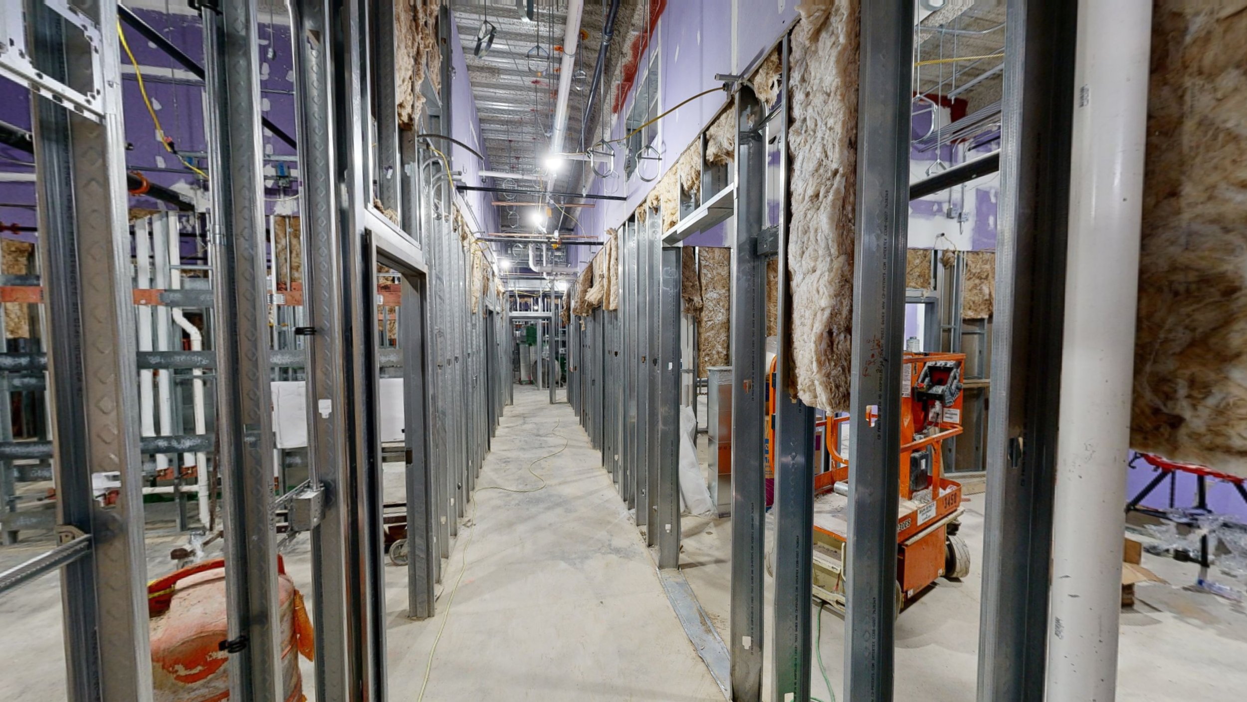 Construction site with metal framing for walls, insulation installed in some areas, electrical wiring visible, and construction equipment and materials present.
