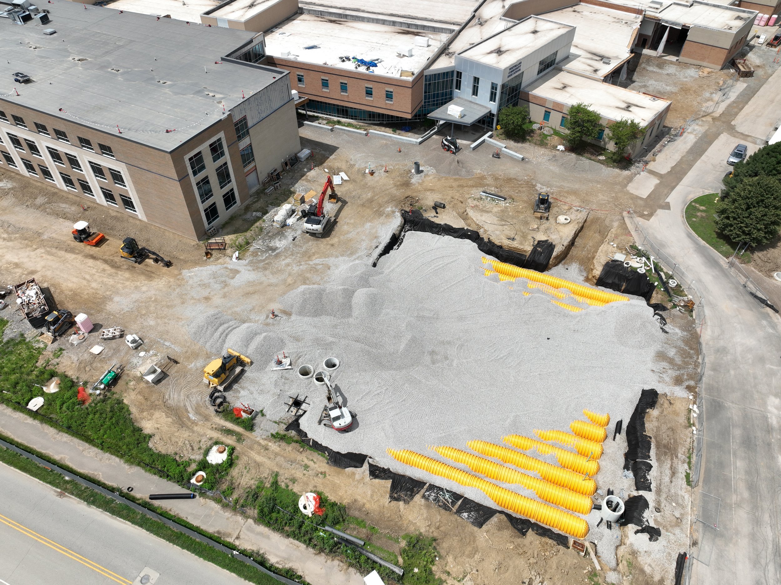 An aerial view of a construction site with a building on the top left and a large gravel area with yellow drainage pipes, construction equipment, and vehicles.