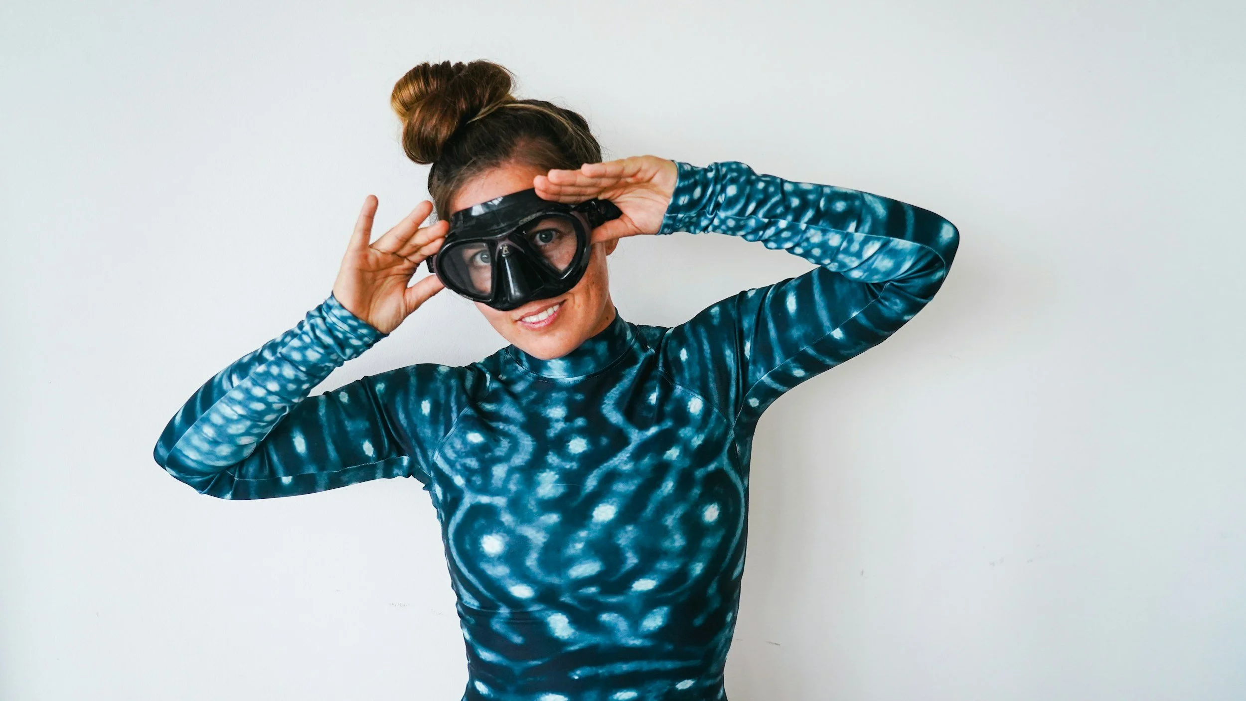 Woman wearing a whale shark patterned shirt standing against a white wall, smiling and adjusting her goggles.