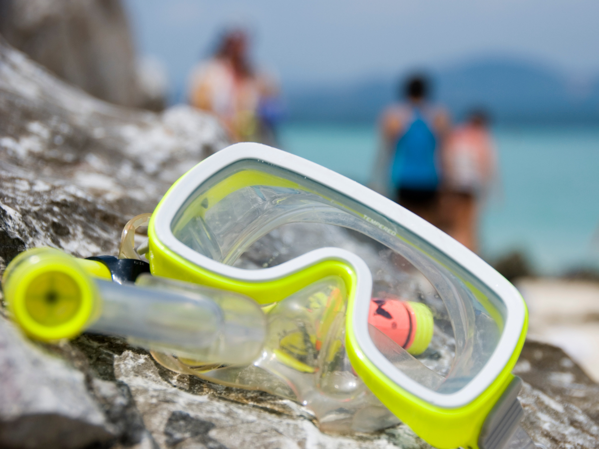 Yellow and clear snorkeling mask and snorkel on a rock with a beach and people in swimsuits in the background