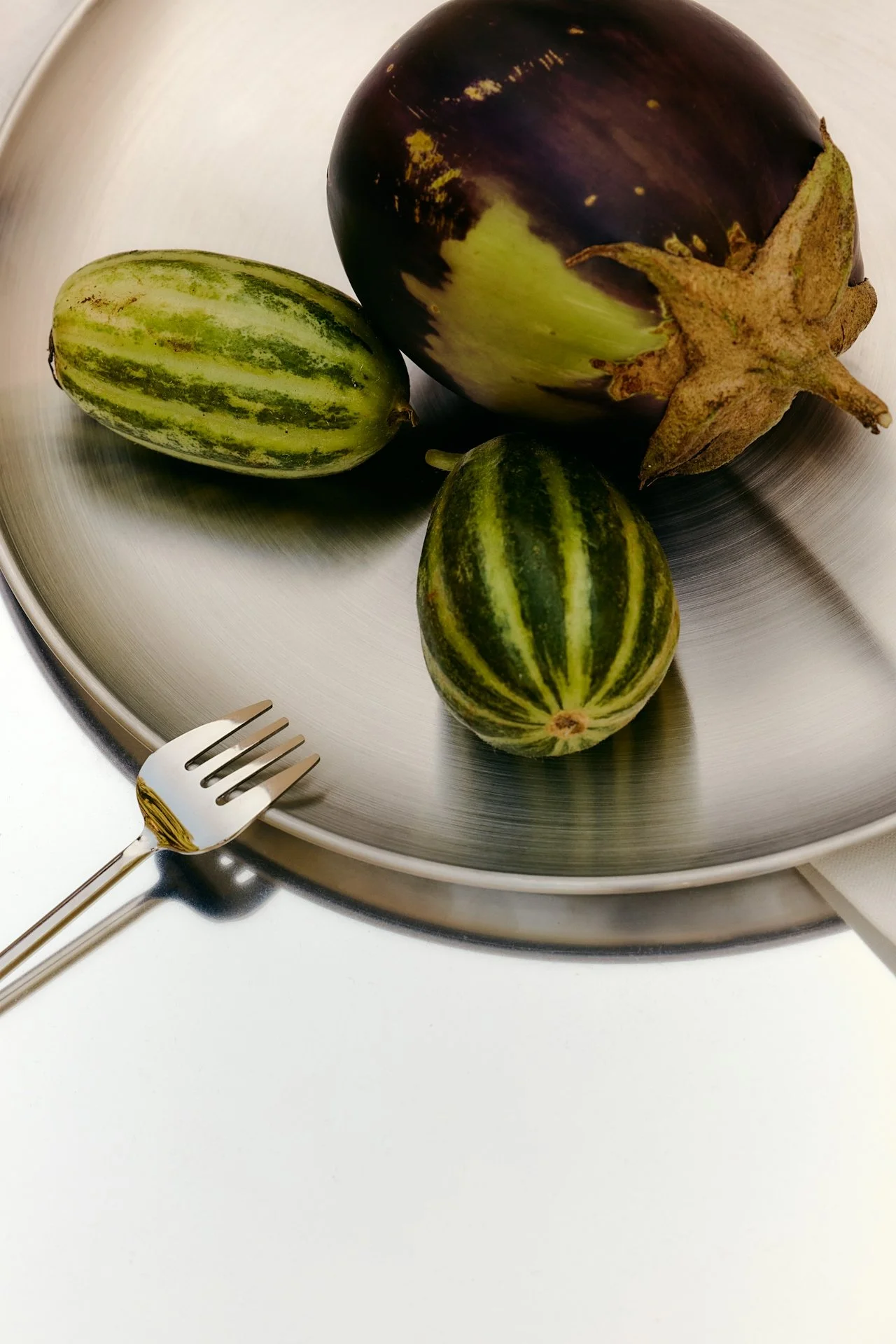 Three small striped eggplants and one larger dark purple eggplant on a silver tray, with a fork resting on the tray edge.