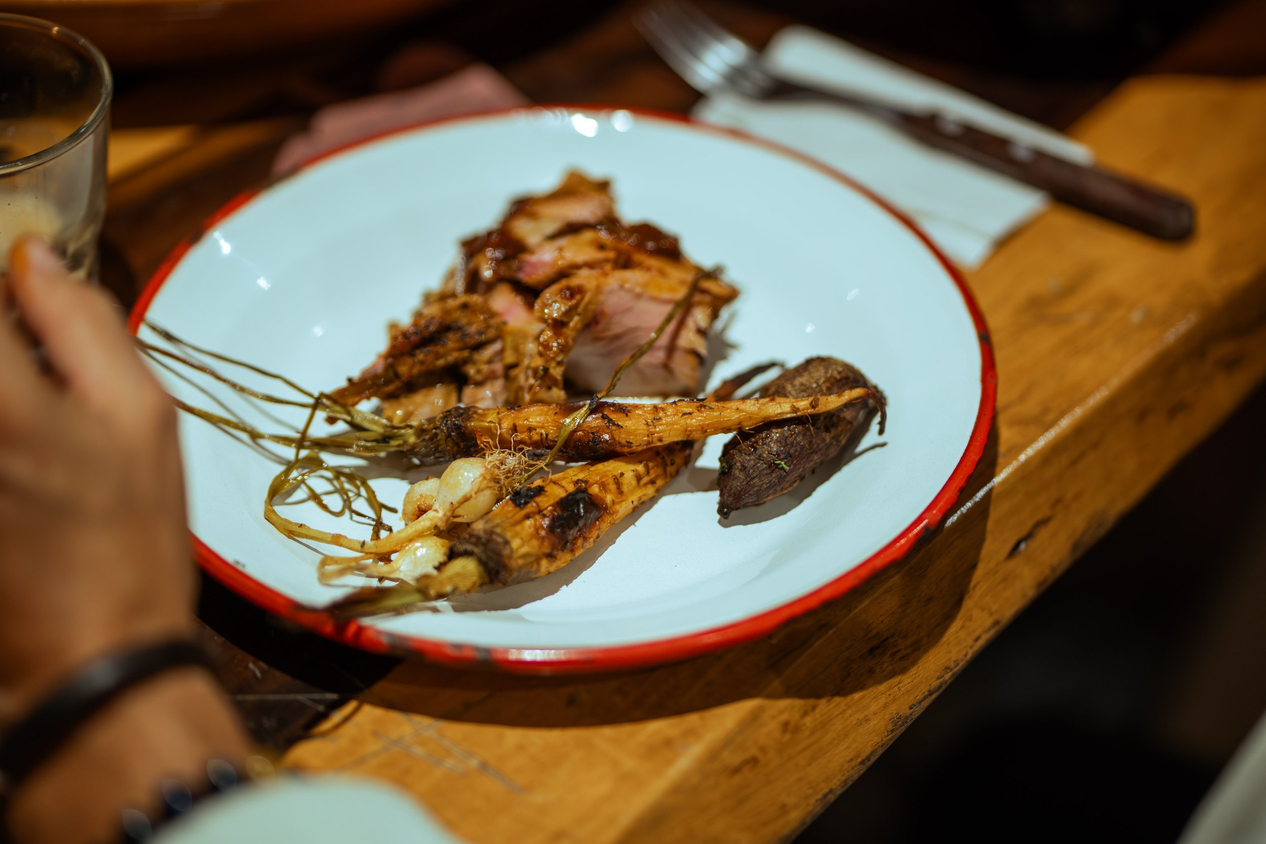 A white plate with a red rim holding grilled meat, carrots, and potatoes on a wooden table.