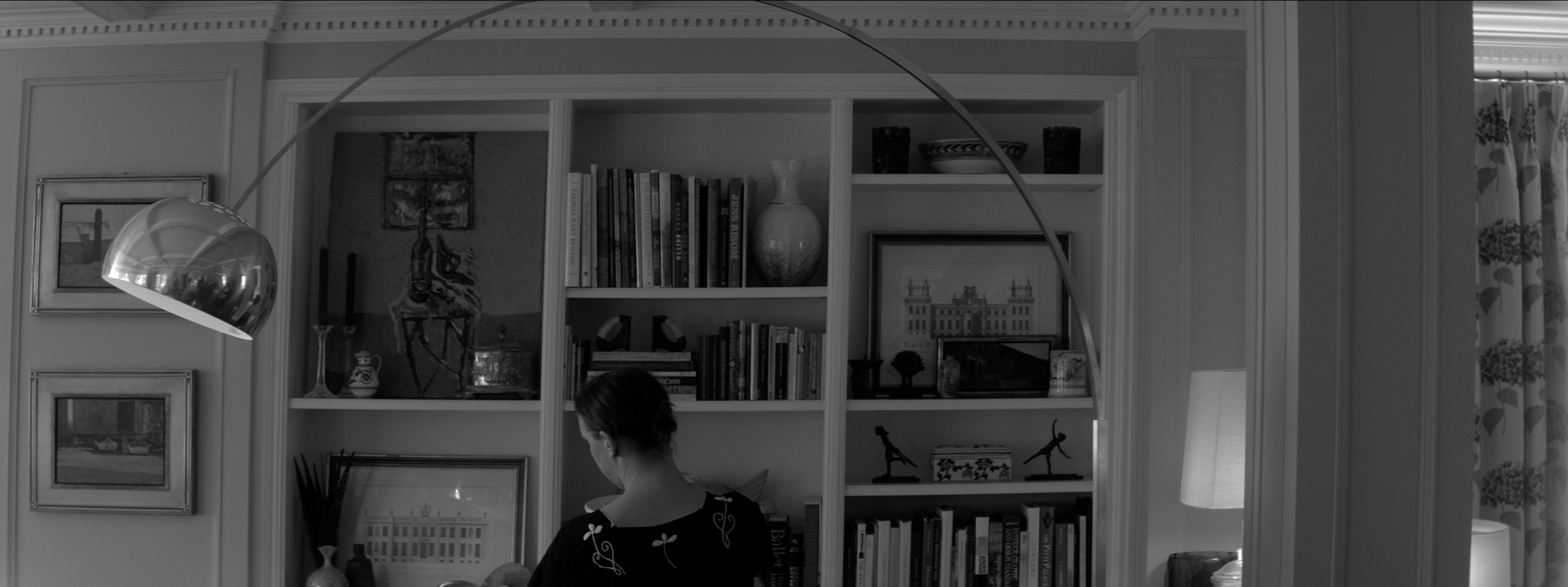 Interior of a room with built-in bookshelf, wall art, and a woman with a bun hairstyle facing away, wearing a black shirt with embroidered designs, standing in front of the bookshelf.