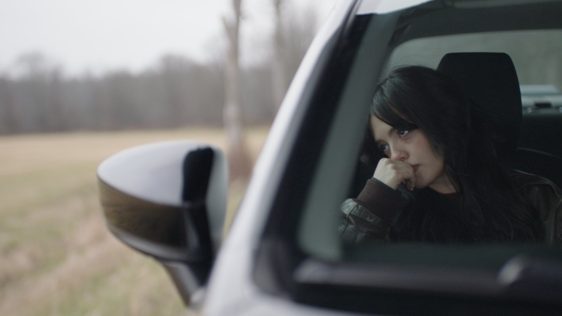 A woman with dark hair sitting inside a car, looking out the window with a thoughtful or concerned expression, with a blurred outdoor background.