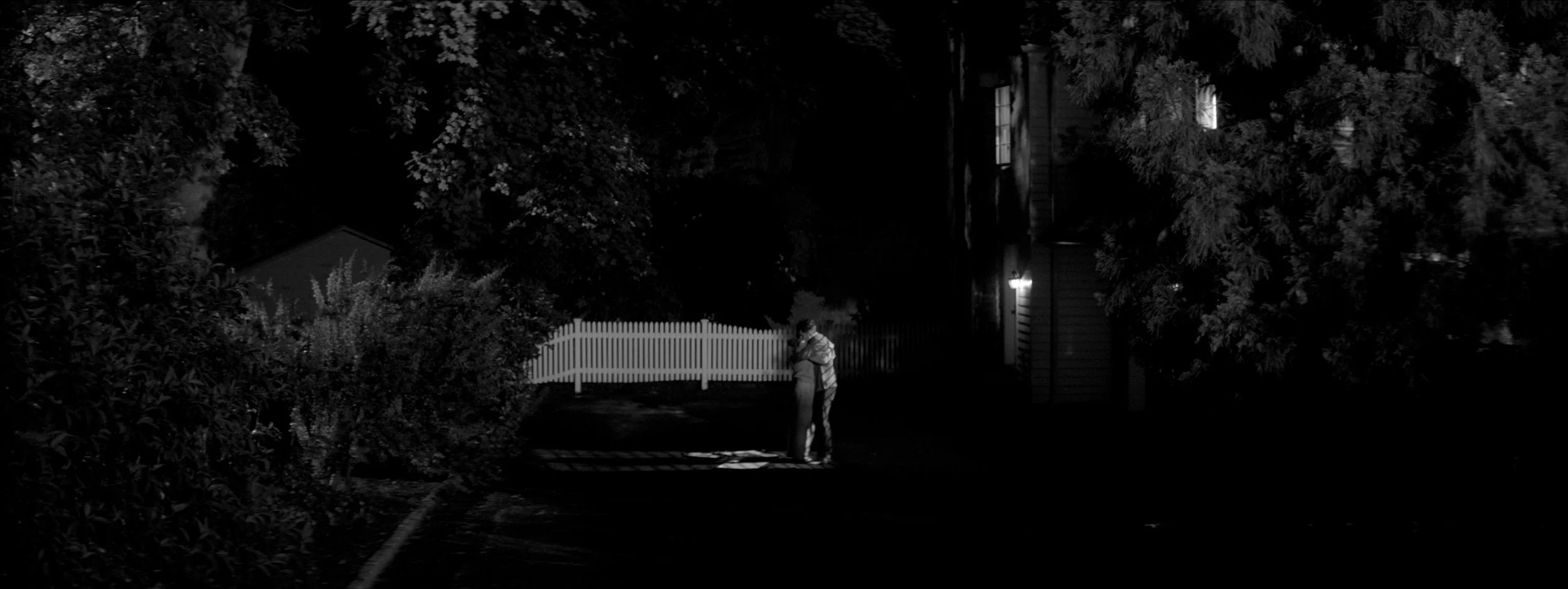 A couple hugging at night outside near a white picket fence, with house windows and trees visible in the dark.