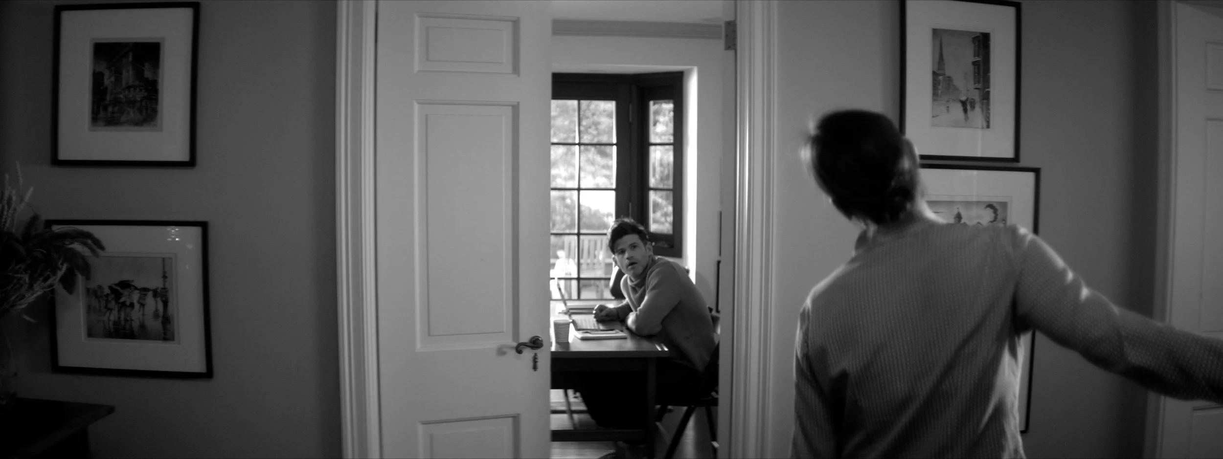 A black and white photo of two people inside a house; one is sitting at a desk in a well-lit room, looking surprised, while the other is standing with their back to the camera, facing the sitting person.