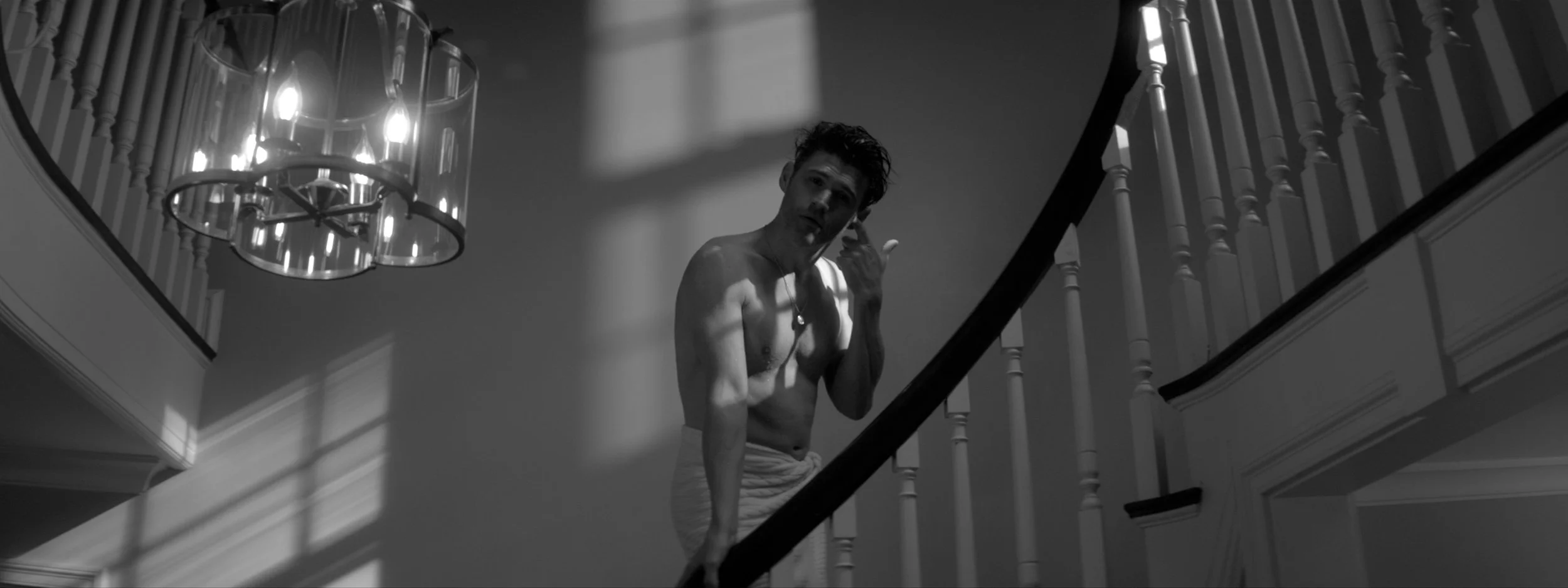 A shirtless man with dark, curly hair looks at the camera while standing on a staircase in a house, with the interior and a chandelier visible in black and white.