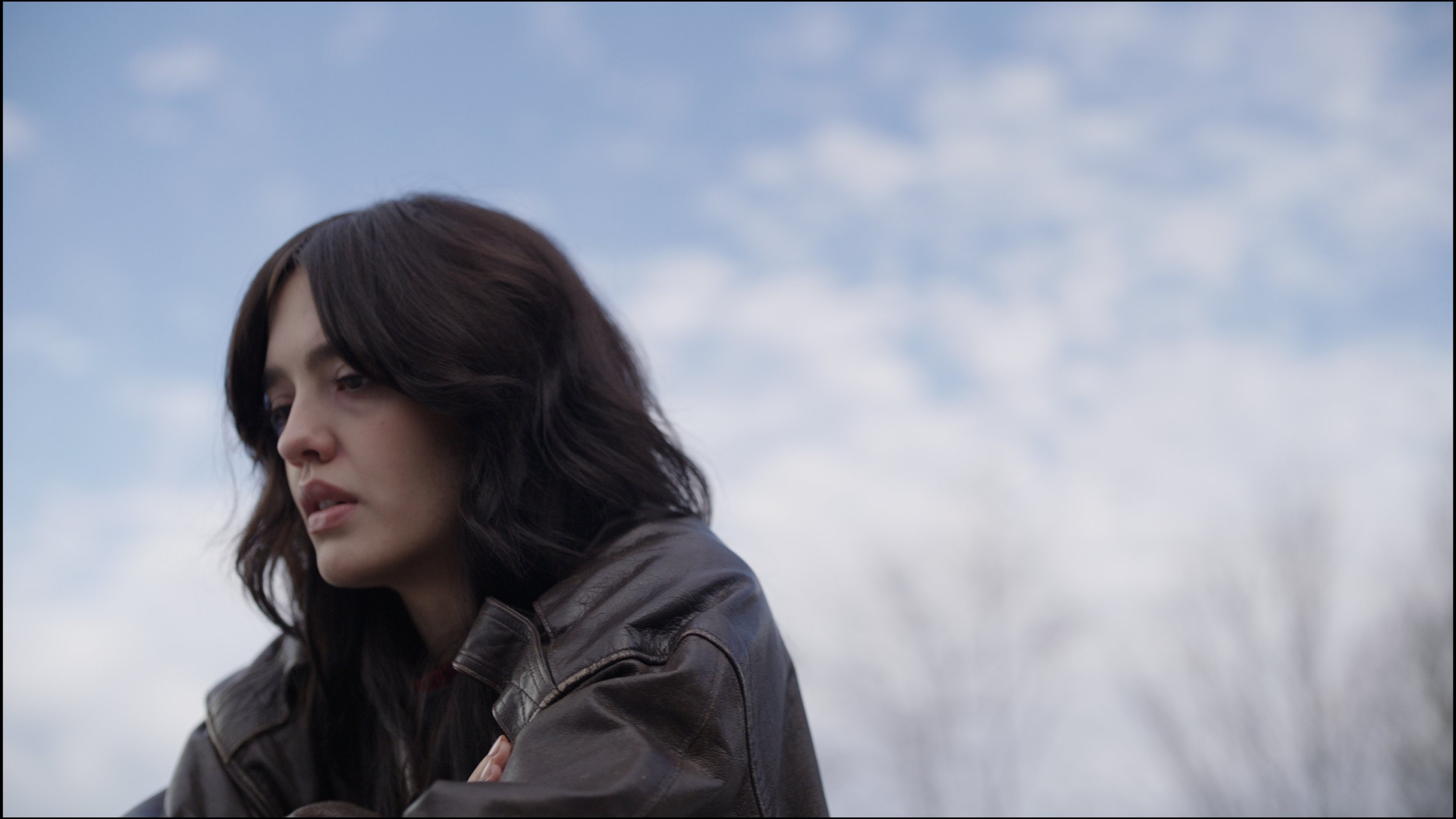 A woman with dark, wavy hair looking thoughtful outdoors, wearing a brown leather jacket against a cloudy sky.
