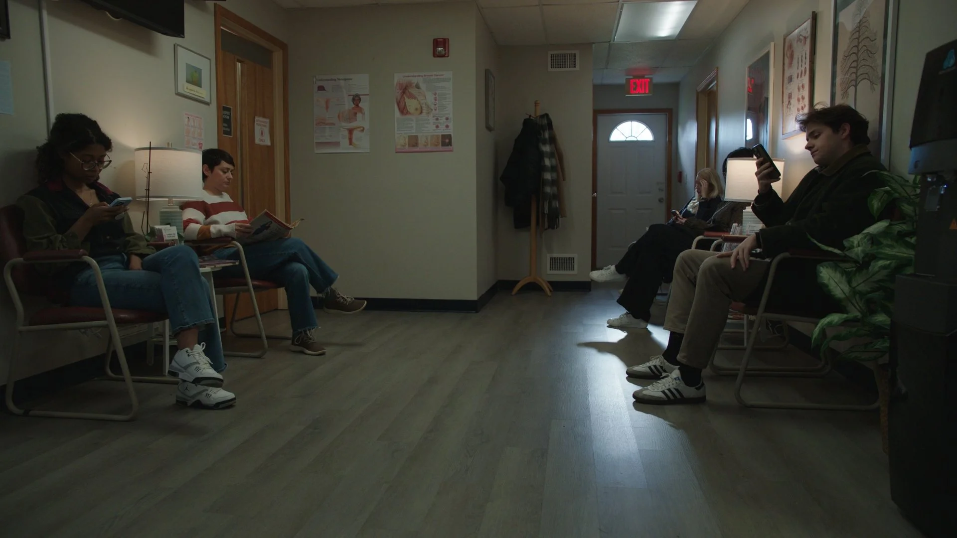 Waiting room with five people seated on chairs, all looking at their phones or reading, posters on the wall, coat rack near the door, and a water cooler on the right.