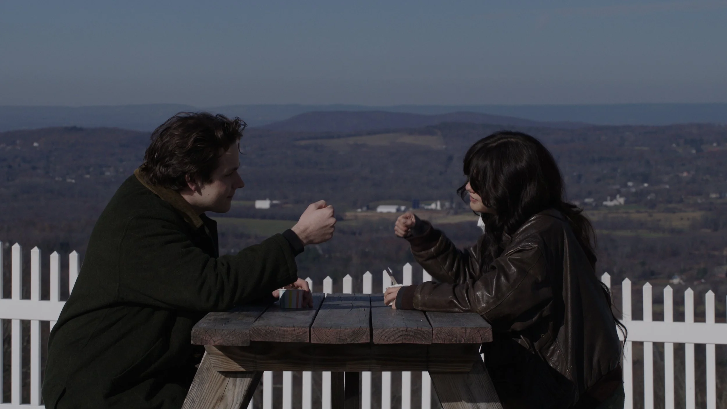 A man and woman sitting across from each other at an outdoor picnic table, engaging in a playful fist bump, with a scenic landscape of hills and a white picket fence in the background.