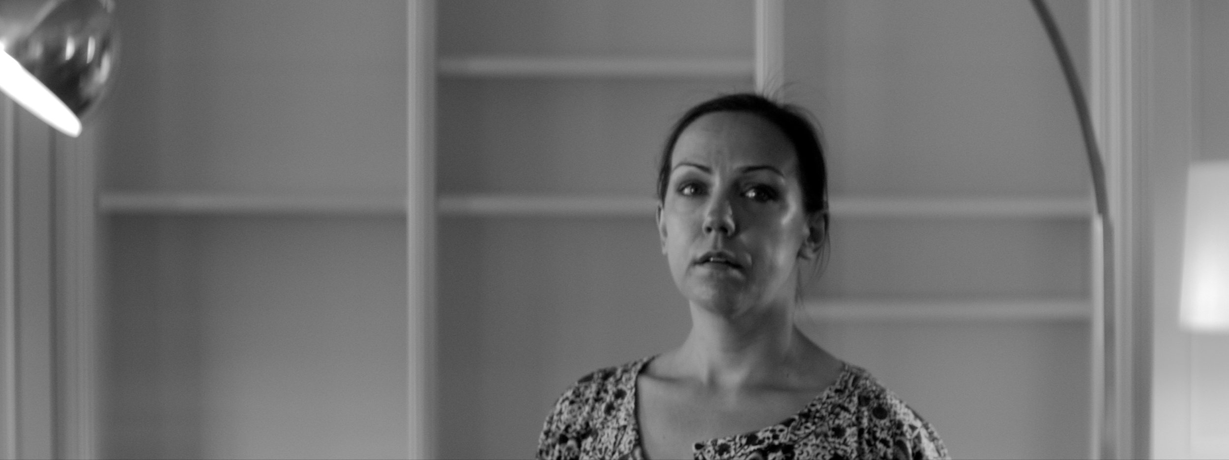 A woman with dark hair looking at her reflection in a mirror, with an empty bookshelf in the background, in black and white.