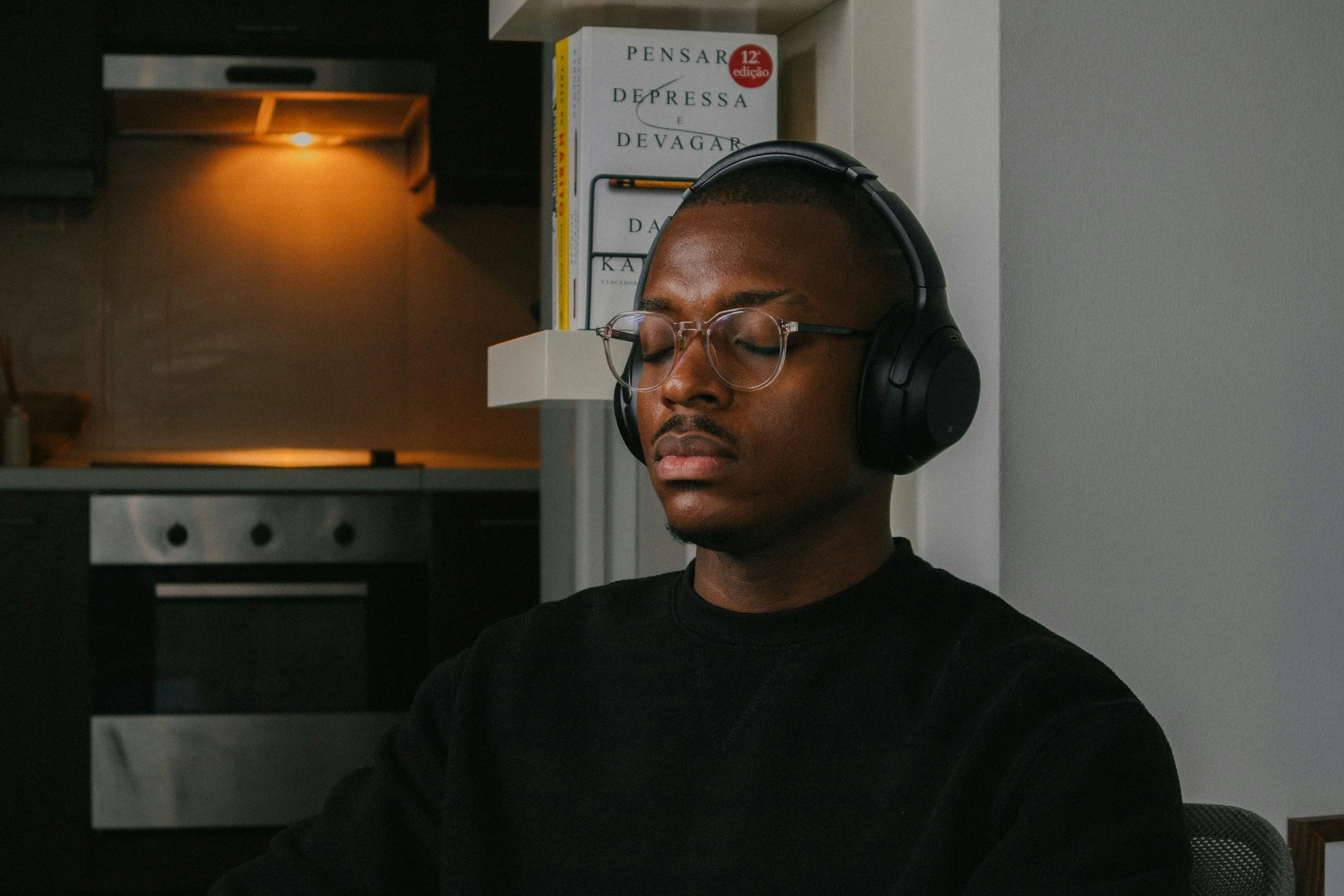 A Man with glasses is sitting in a dimly lit room. He is wearing headphones and has his eyes closed while he meditates.