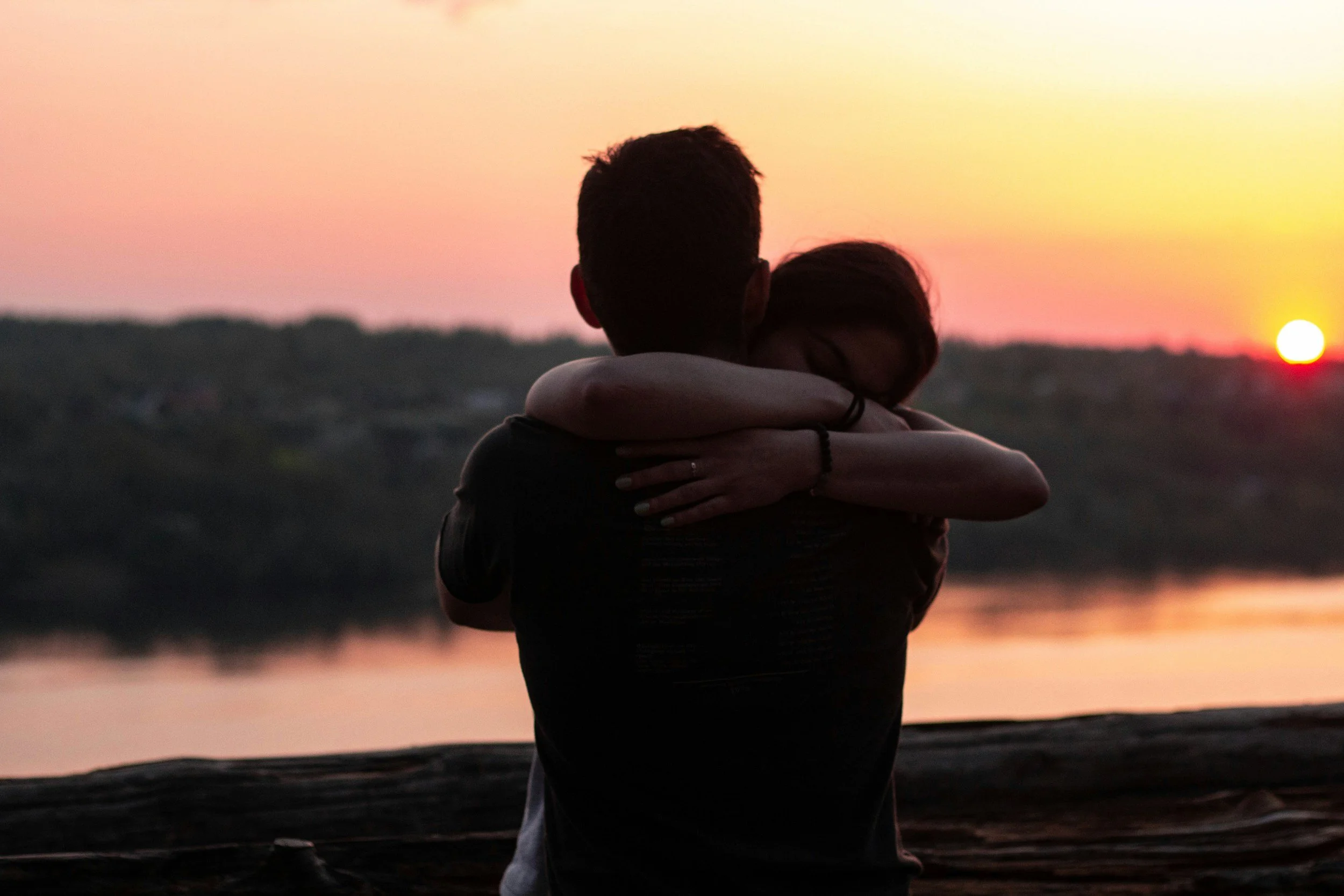 Image shows a dark silhouette of a man and woman hugging in the foreground. In the background there is a body of water and trees blurred. The sun is setting which casts a calming orange gradient throughout the sky, which is reflected in the water.