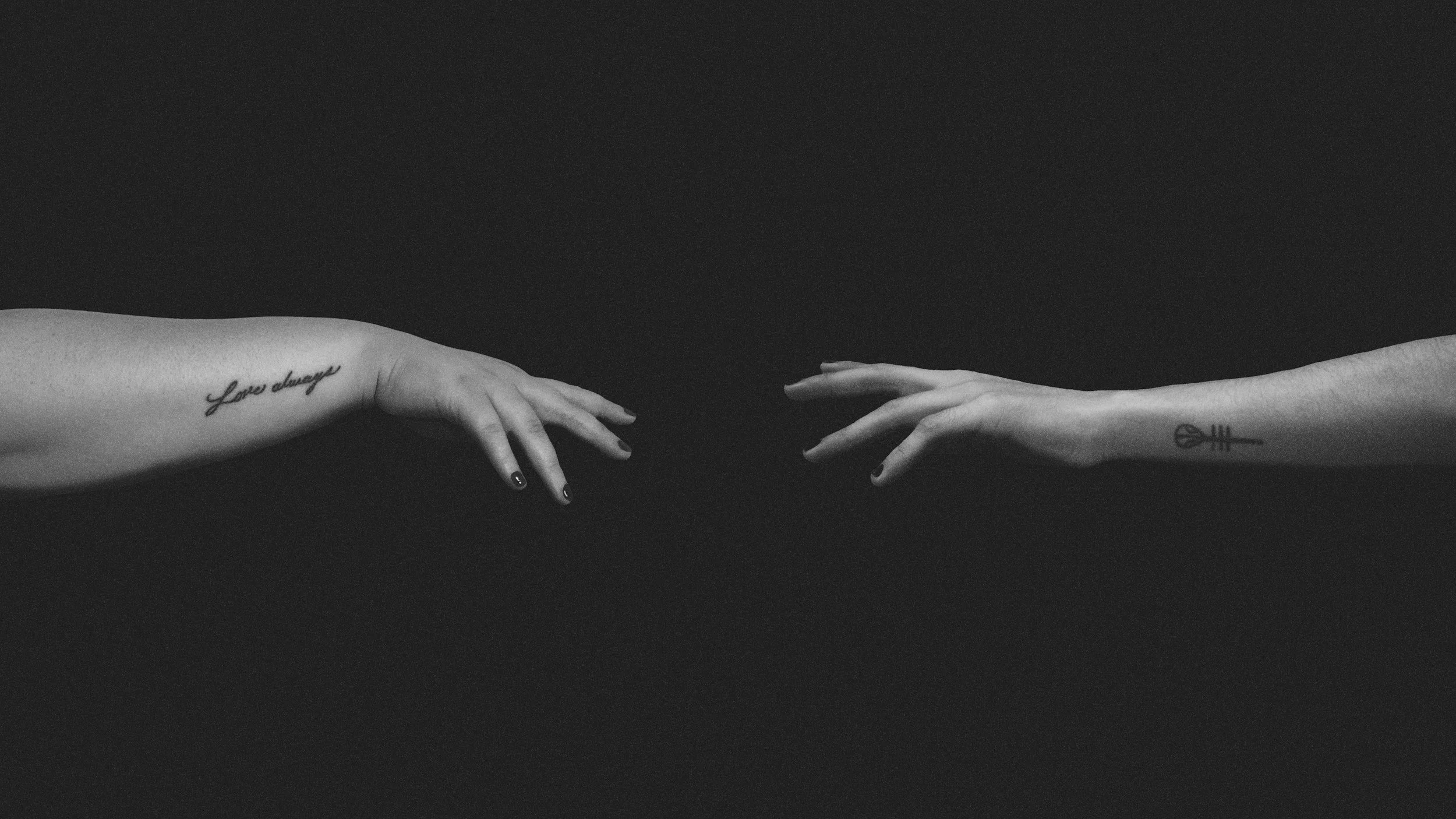 Image has a black background. Two arms are reaching for each other, each arm belonging to a different person. One arm enters from the left side, reaching to the middle. The other arm enters from the right, reaching to the middle. The hands dont touch